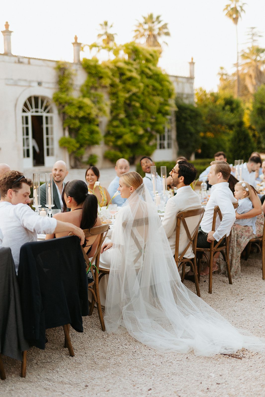 Elegant outdoor wedding reception with flowy white veil and guests seated at rustic tables against historic white building.