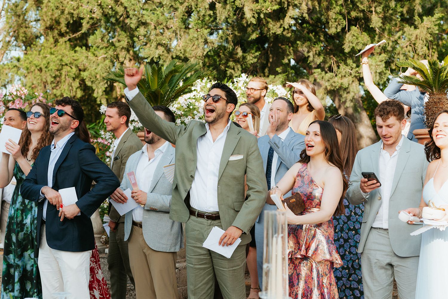 Wedding guests in summer attire raise their glasses and celebrate during an outdoor reception under trees.