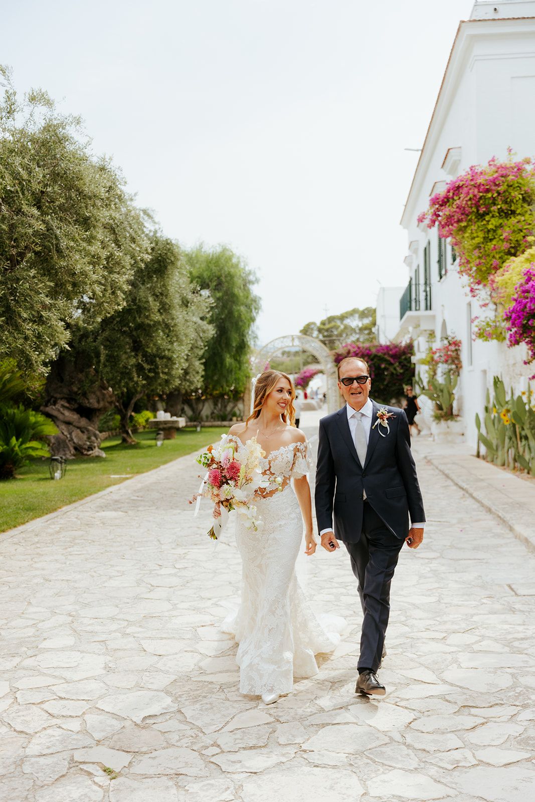 A couple walks along a stone pathway with white walls and colorful bougainvillea flowers in a wedding celebration setting.