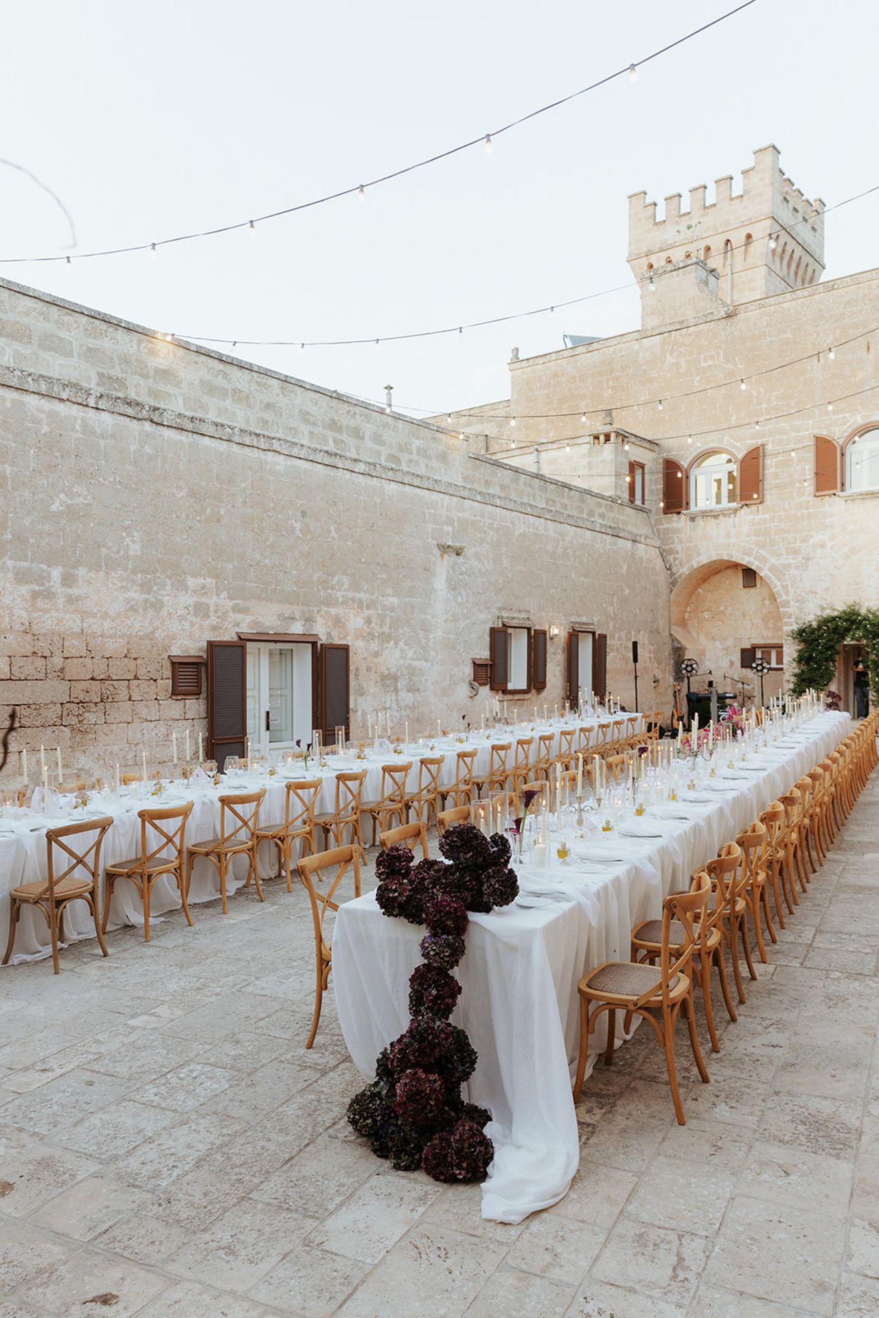 Long elegant wedding reception table setup in a historic courtyard with stone walls and wooden chairs.