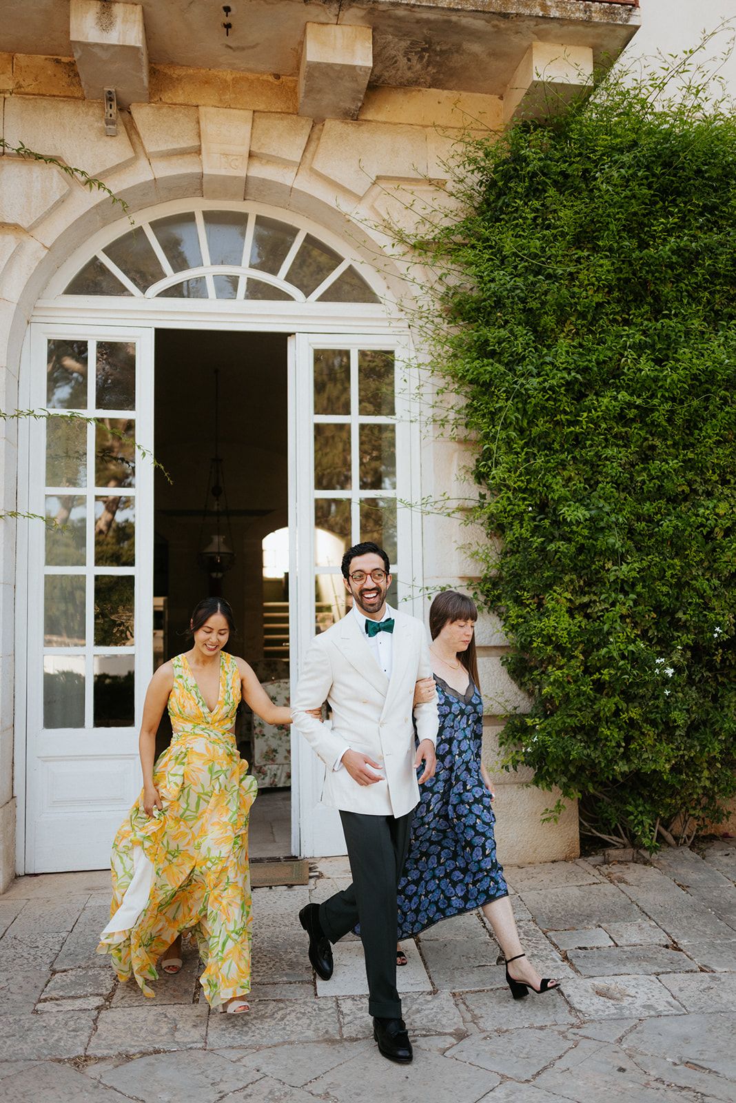 People walking out of an elegant stone entrance with arched doorway and lush greenery at a formal event.
