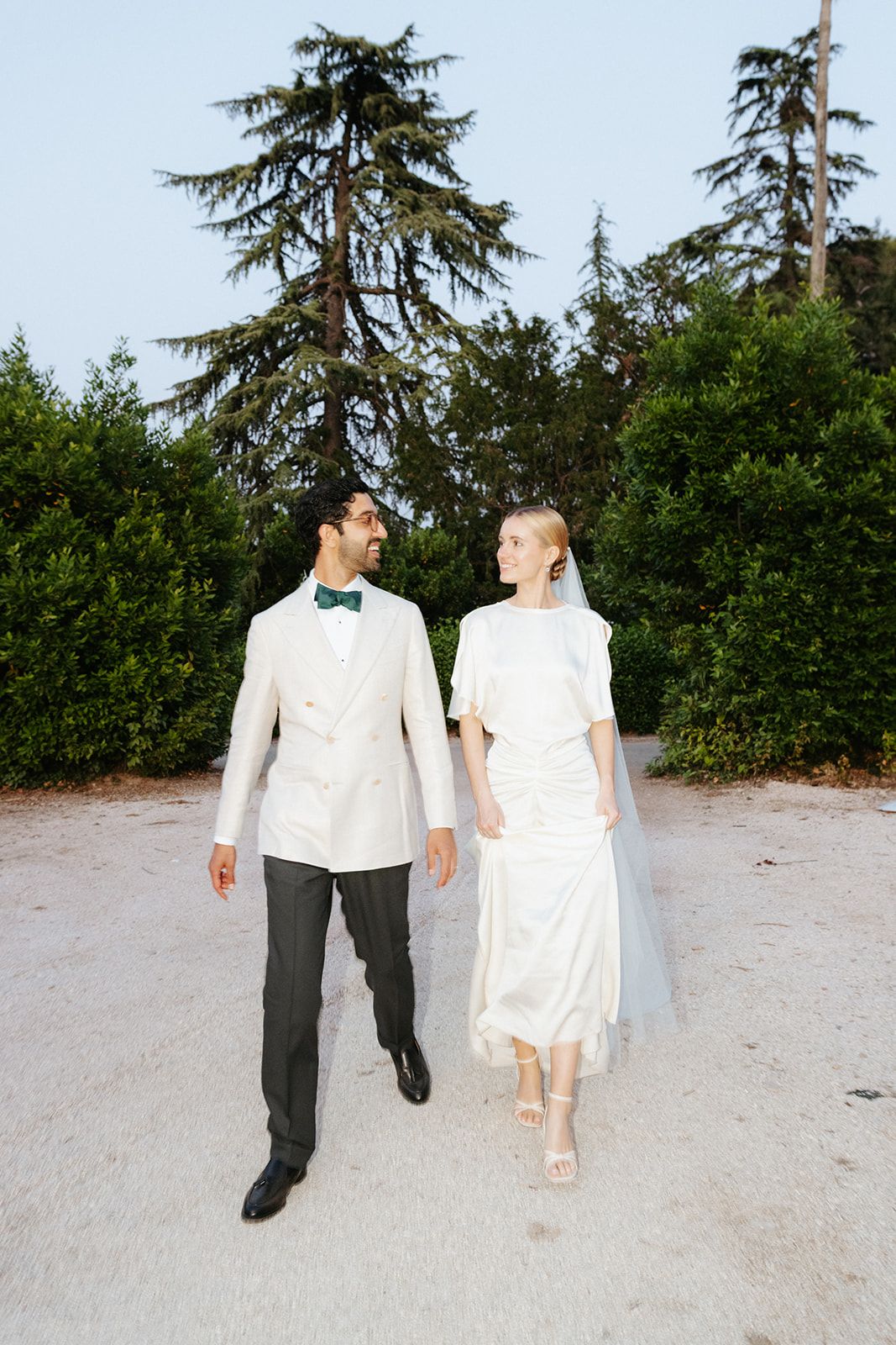 A couple in wedding attire stroll hand in hand along a sandy beach with evergreen trees in the background.