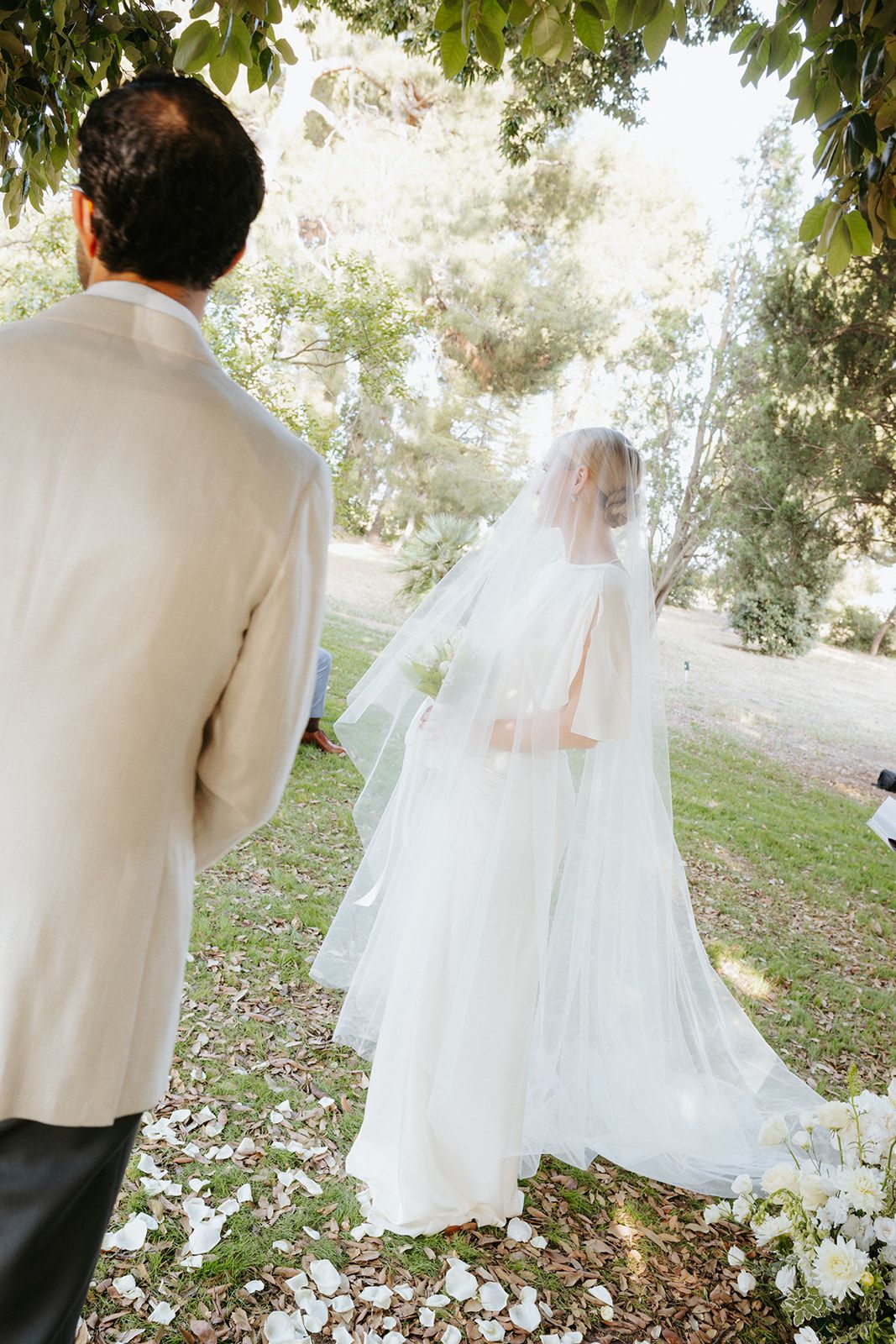 A bride in a flowing white gown and veil walks ahead on grass while someone in a white suit jacket watches from behind.