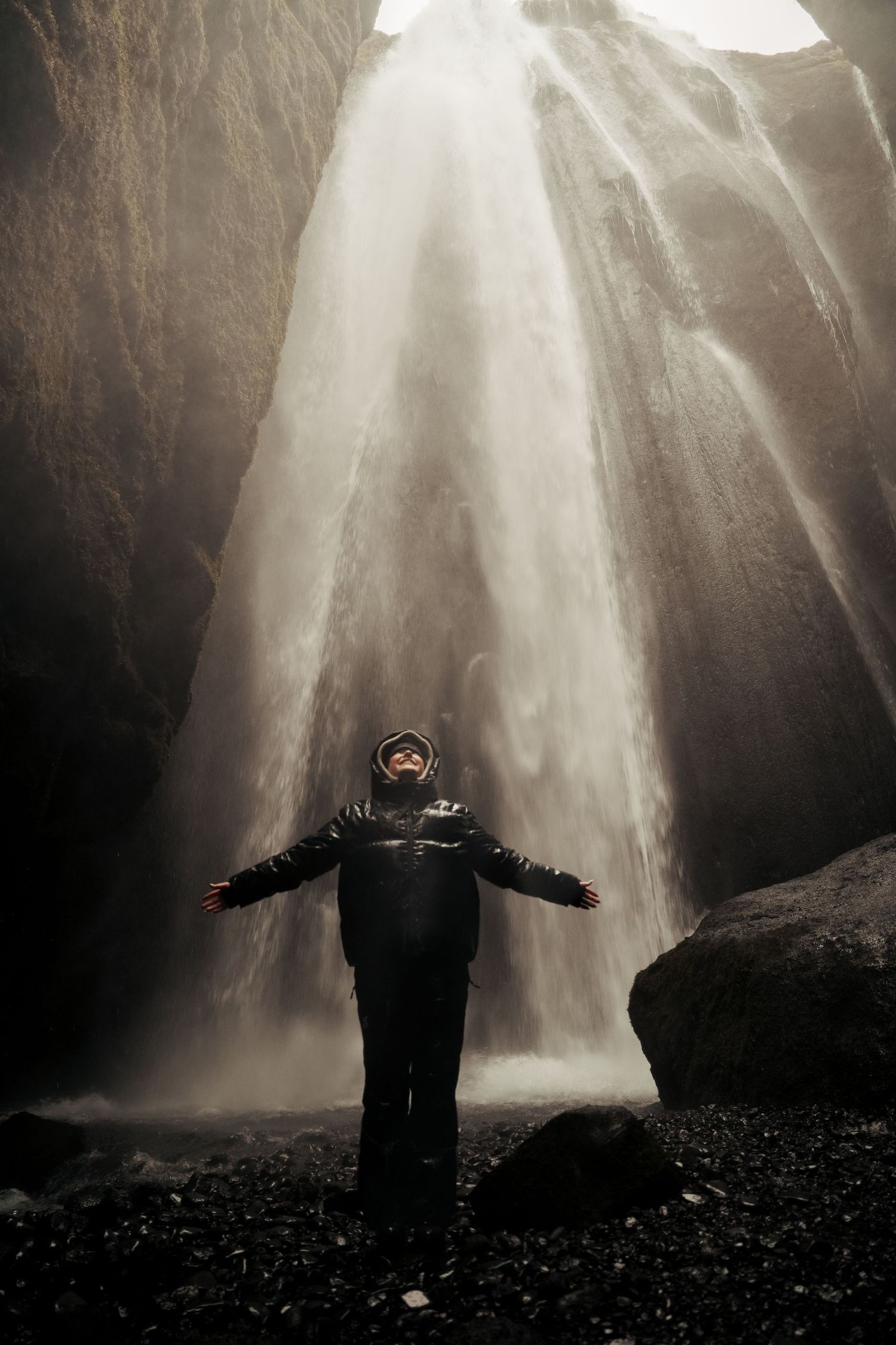 Silhouetted figure stands with arms outstretched beneath a majestic waterfall in a dark cave. Iceland.