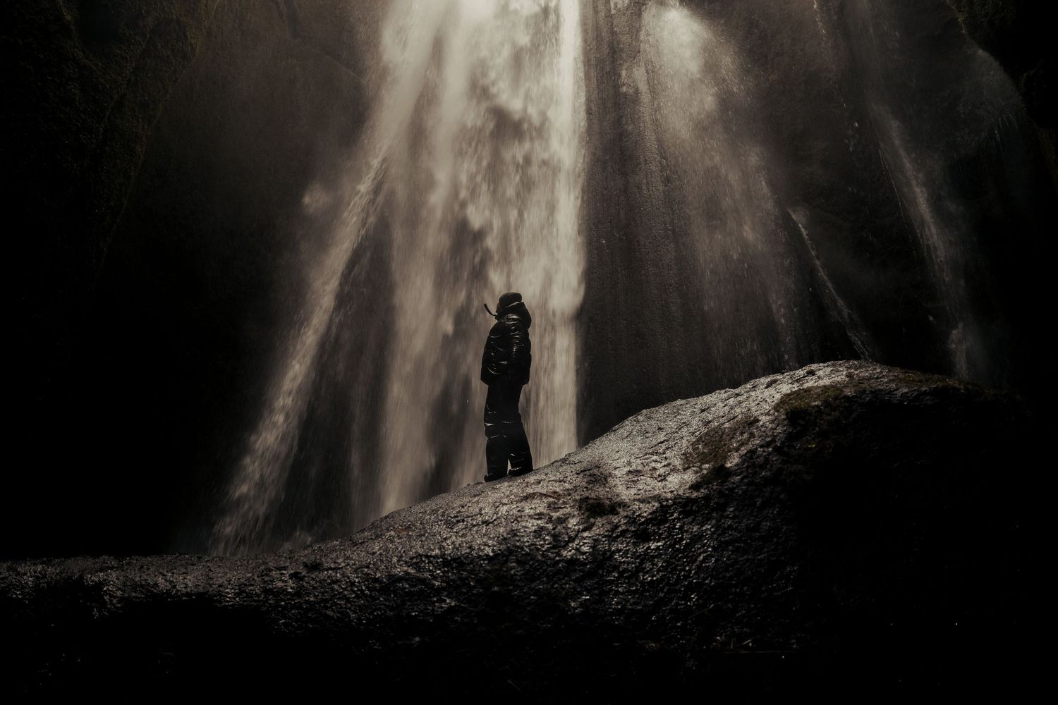 Dramatic waterfall cascades down in a dark cave with a small silhouetted figure visible below. Iceland.