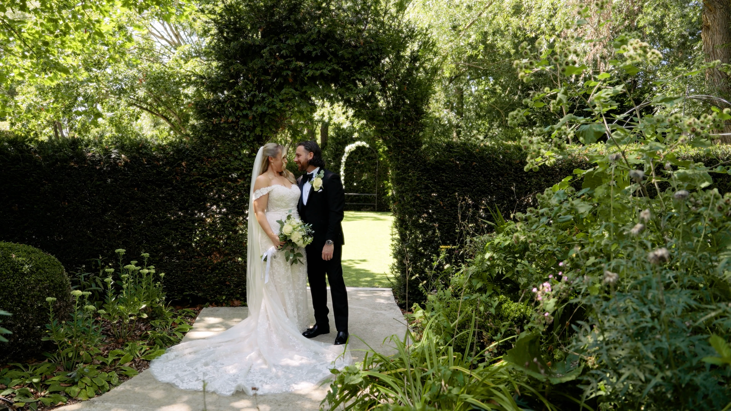 A wedding ceremony takes place in a lush garden setting with natural greenery and sunlight filtering through the trees. The Barn Brasserie.