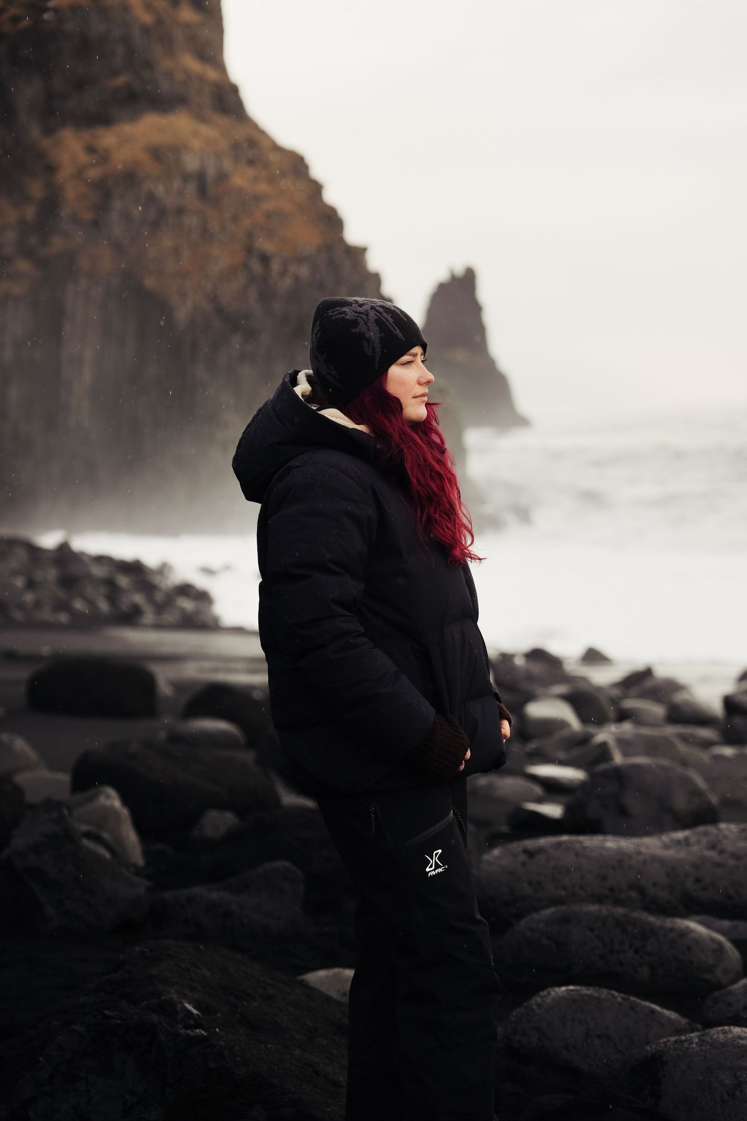 A person in a black coat stands on volcanic rocks with dramatic coastal cliffs in the background. Iceland.