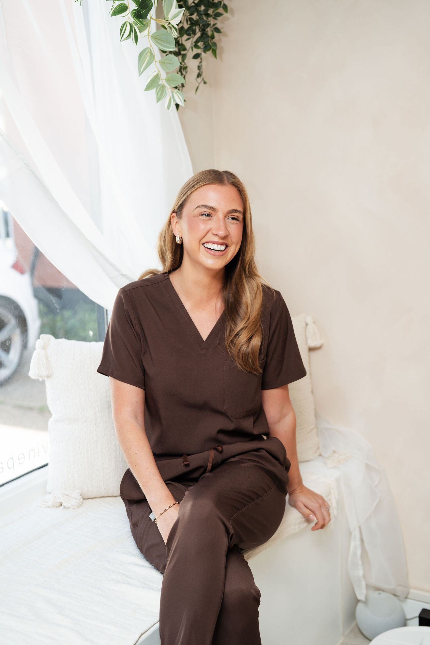 Person in brown scrubs sitting casually by window with natural light and plants. Colchester, Essex.