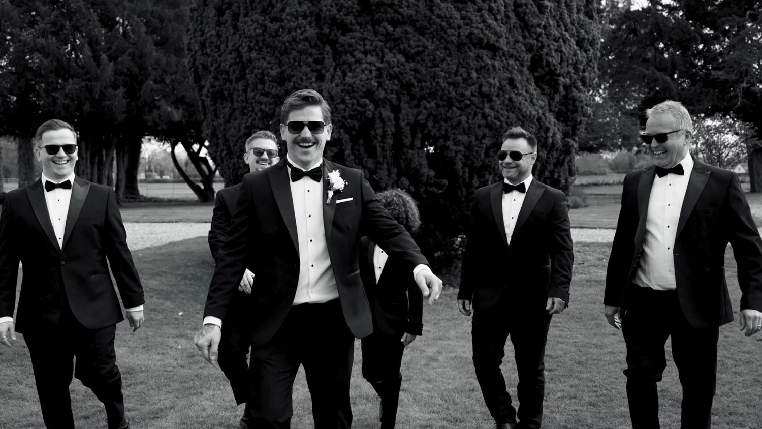 Groomsmen in formal attire walking together on grassy path at sunset. Gosfield Hall.