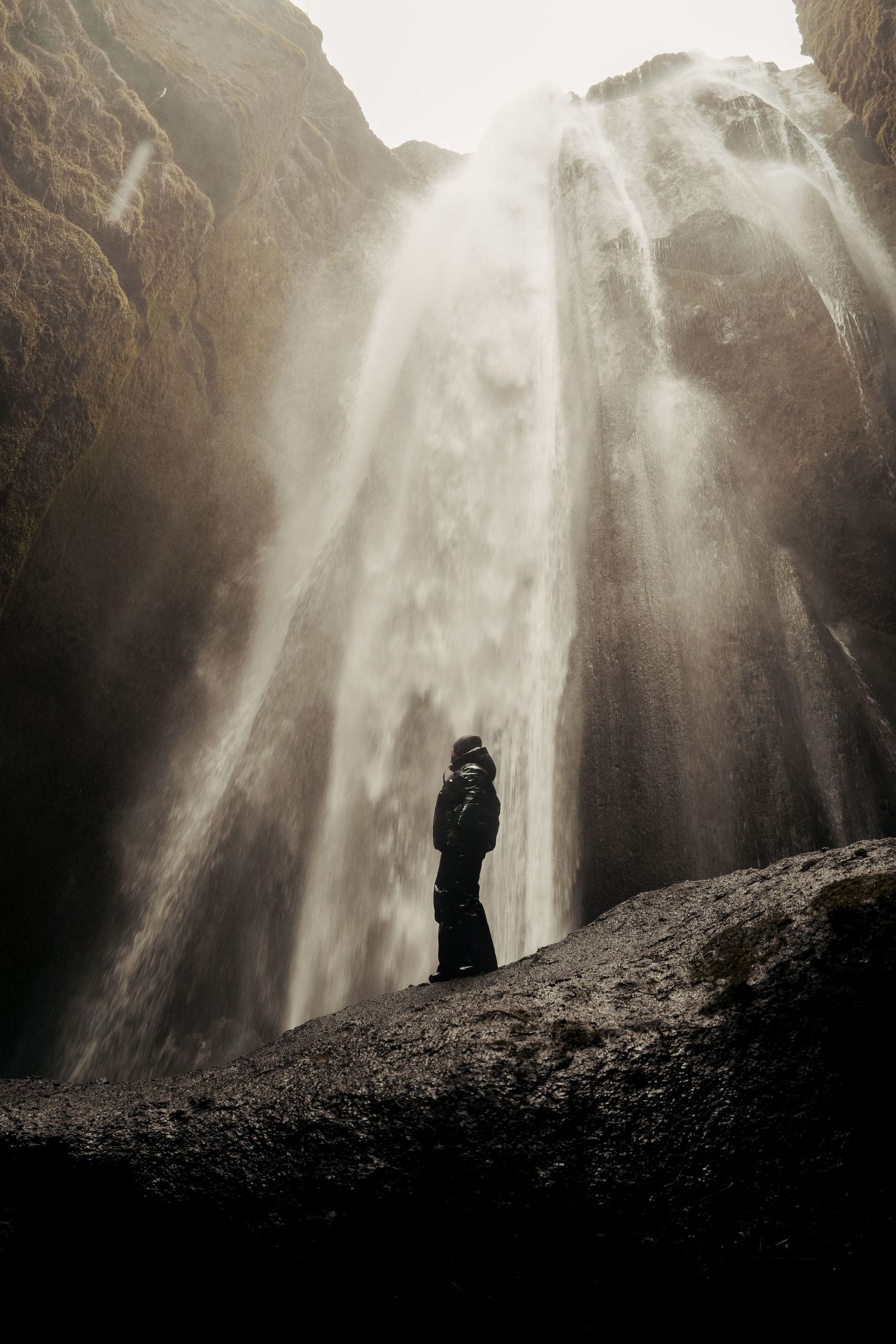 Silhouetted figure stands beneath towering misty waterfall in monochrome scene. Iceland.