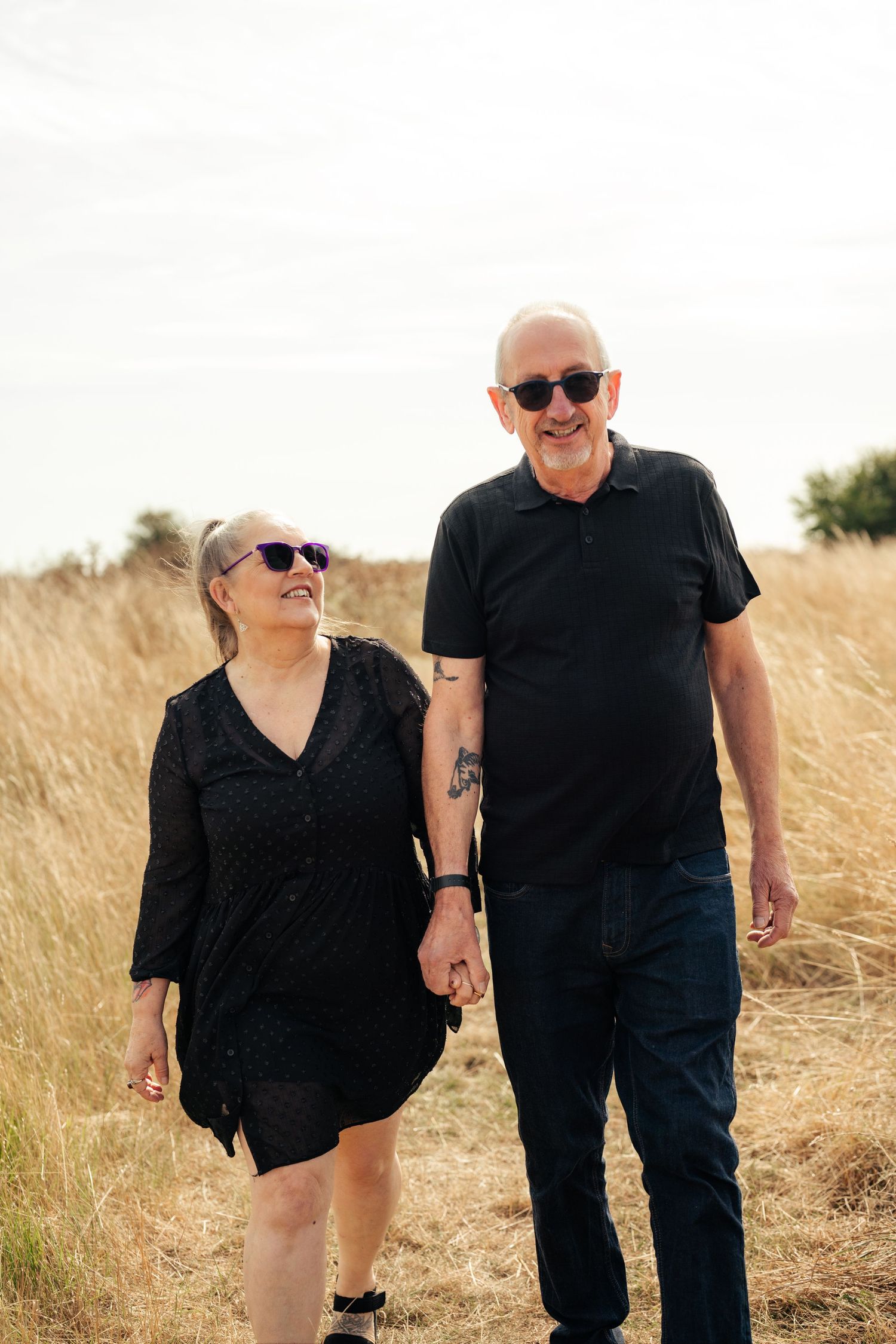 A couple walks together through a golden field wearing matching black outfits. Earlhams Beach, Harwich, Essex.