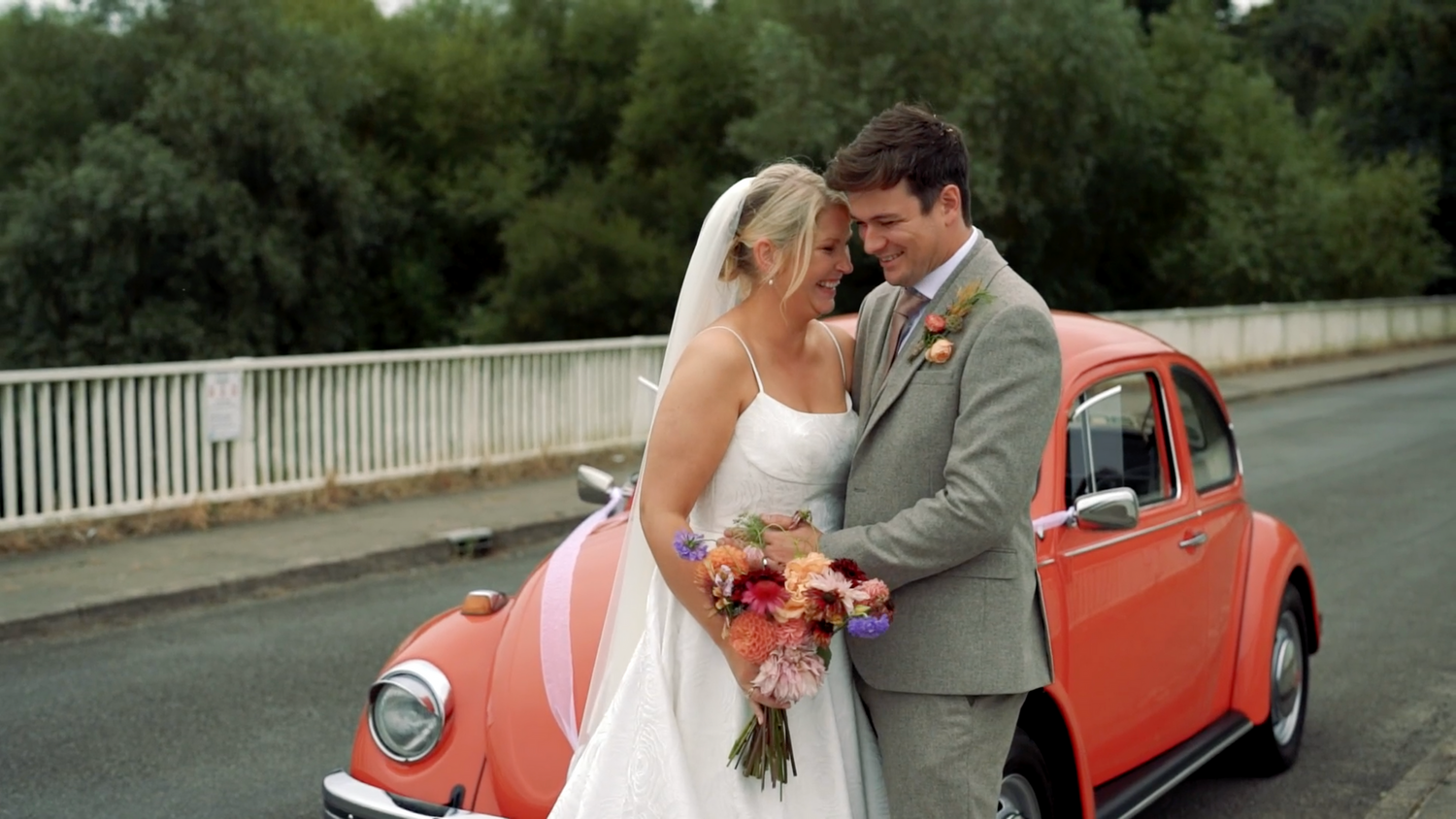 Newlywed couple poses romantically by vintage coral-colored car on their wedding day. Suffolk, Uk.