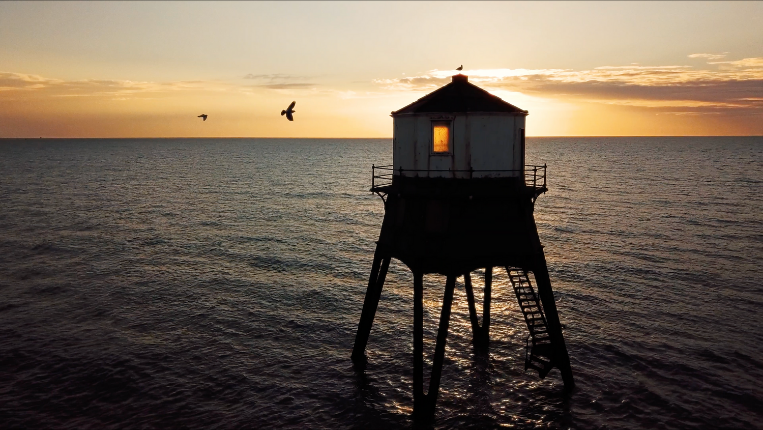 Silhouette of stilted beach house against dramatic ocean sunset with birds flying. Dovercourt, Essex.