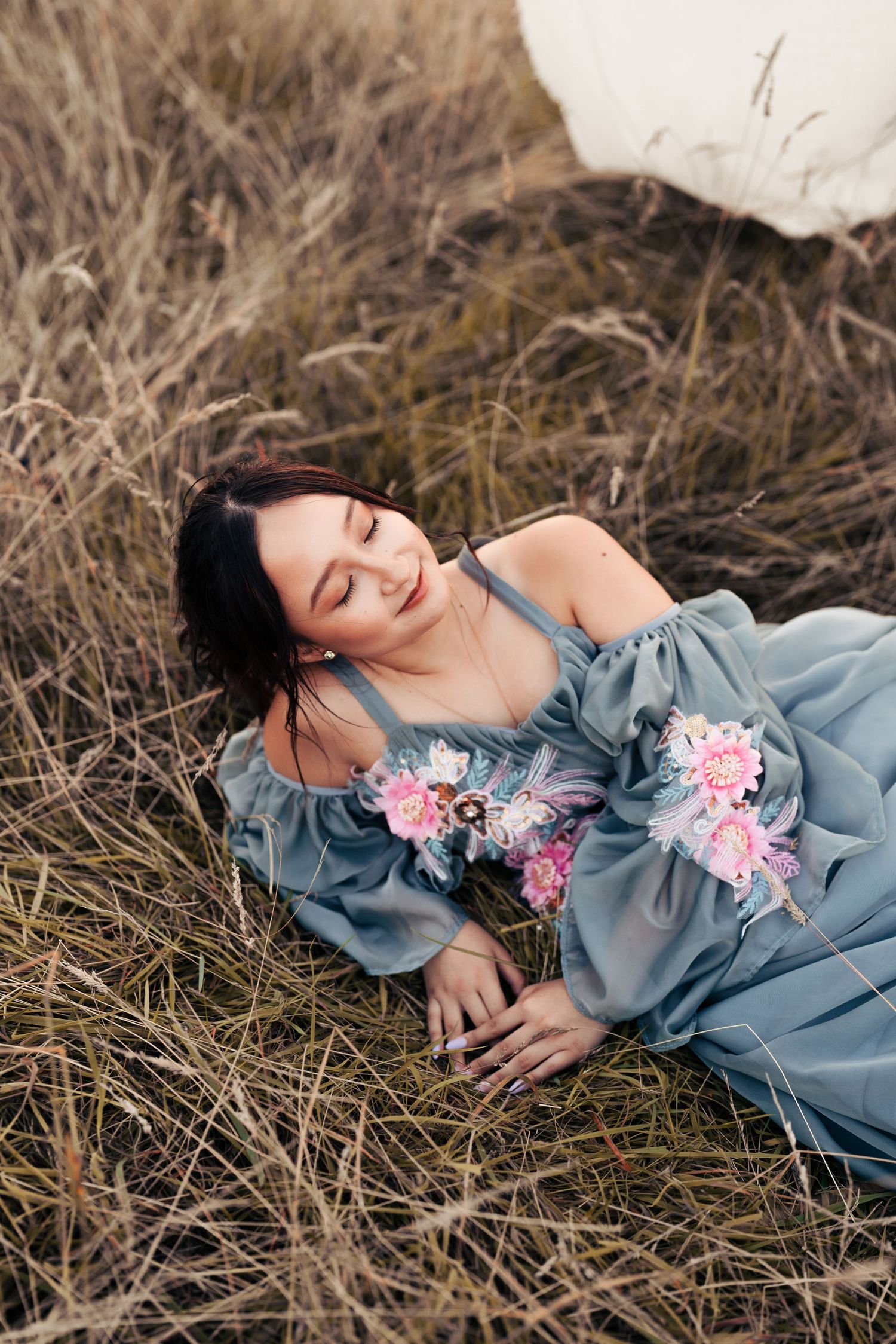 Artistic portrait series showing someone laying in dry grass wearing a romantic blue dress with floral accents. Wrabness, Essex.