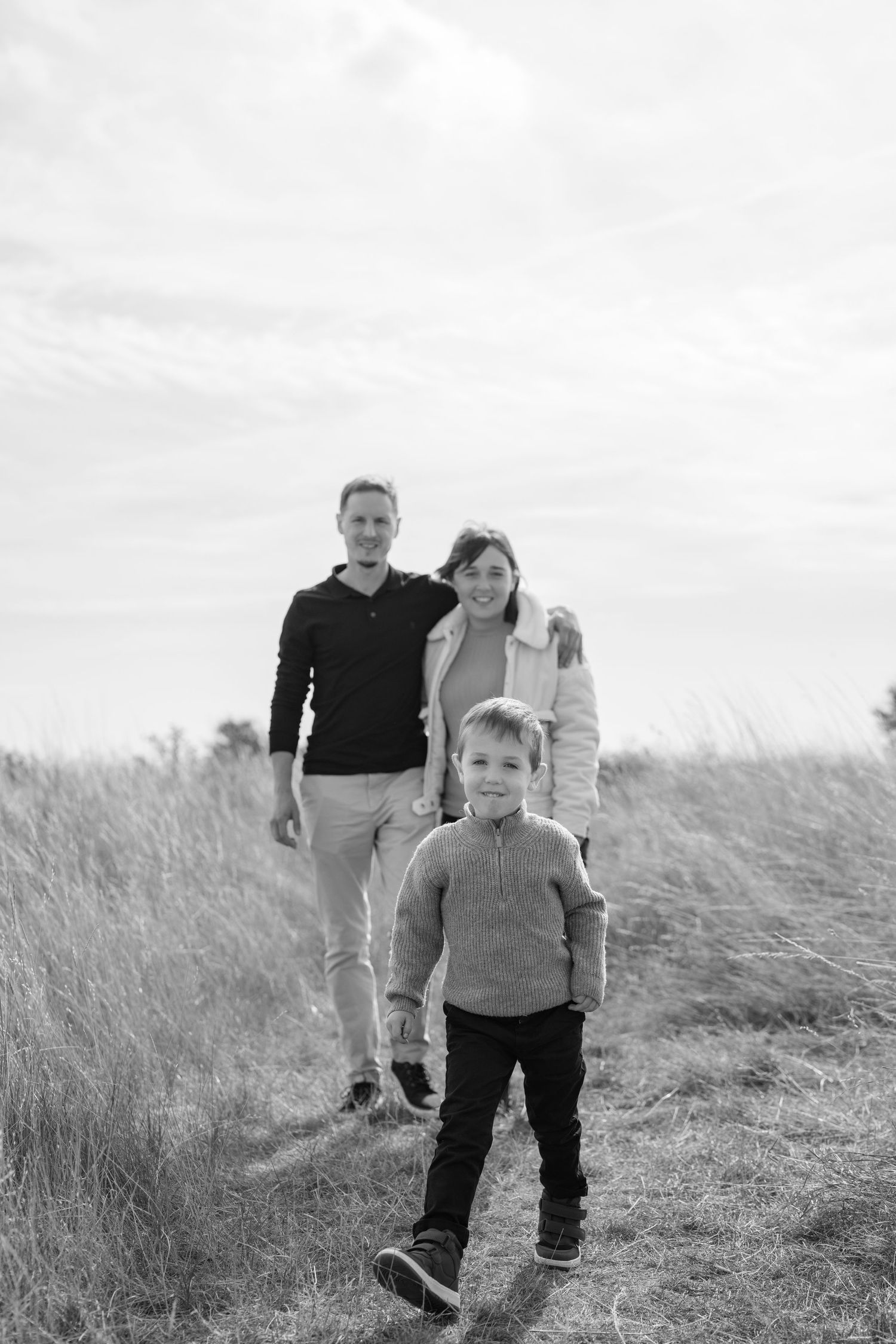 A family walks together through tall grass on a cloudy day during an outdoor photoshoot. Earlhams Beach, Harwich, Essex.
