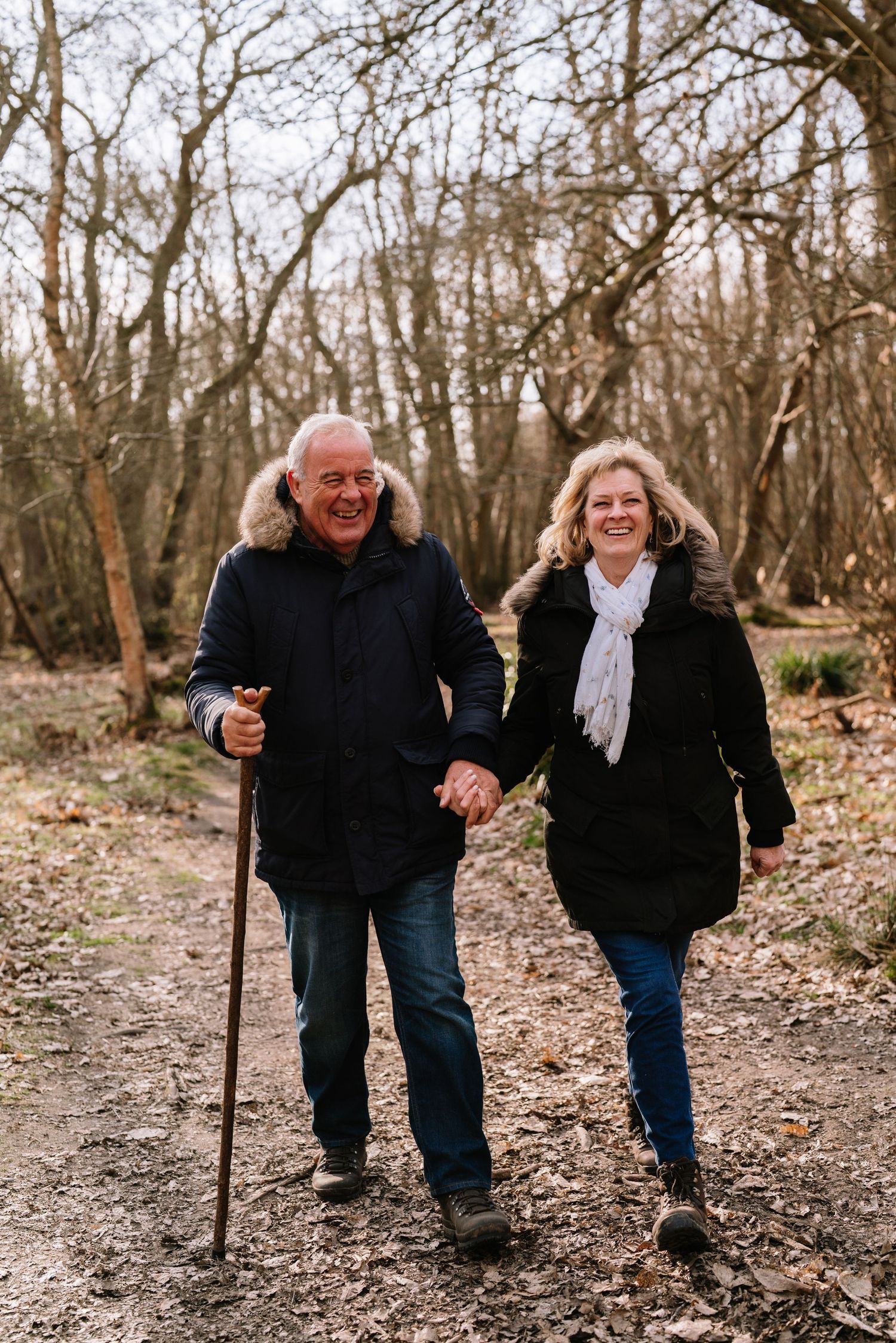 Couple walking together with walking sticks on a forest path covered in fallen leaves. Wrabness Woods, Essex.