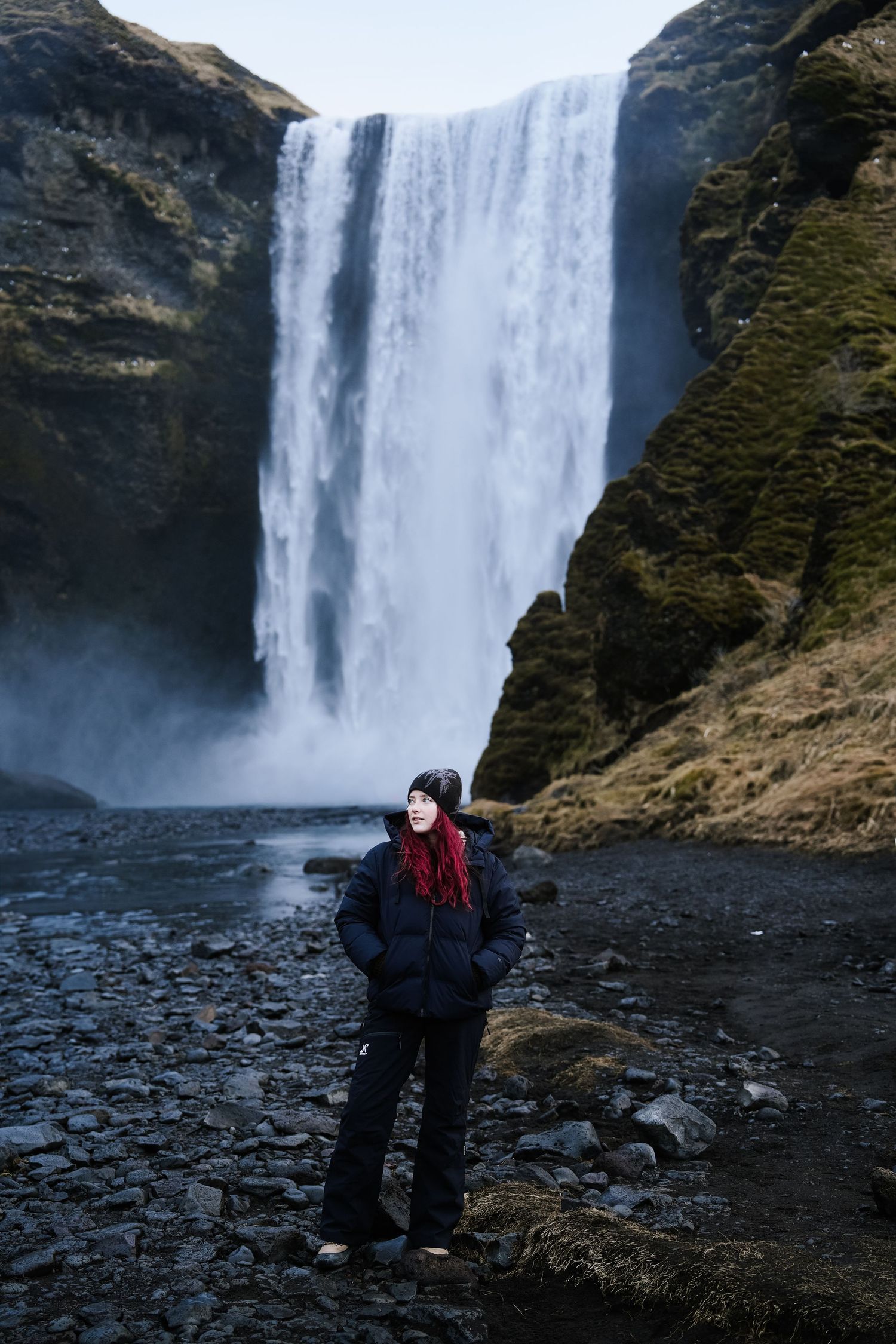 A figure stands on rocky terrain beneath a massive waterfall in Iceland's rugged landscape.