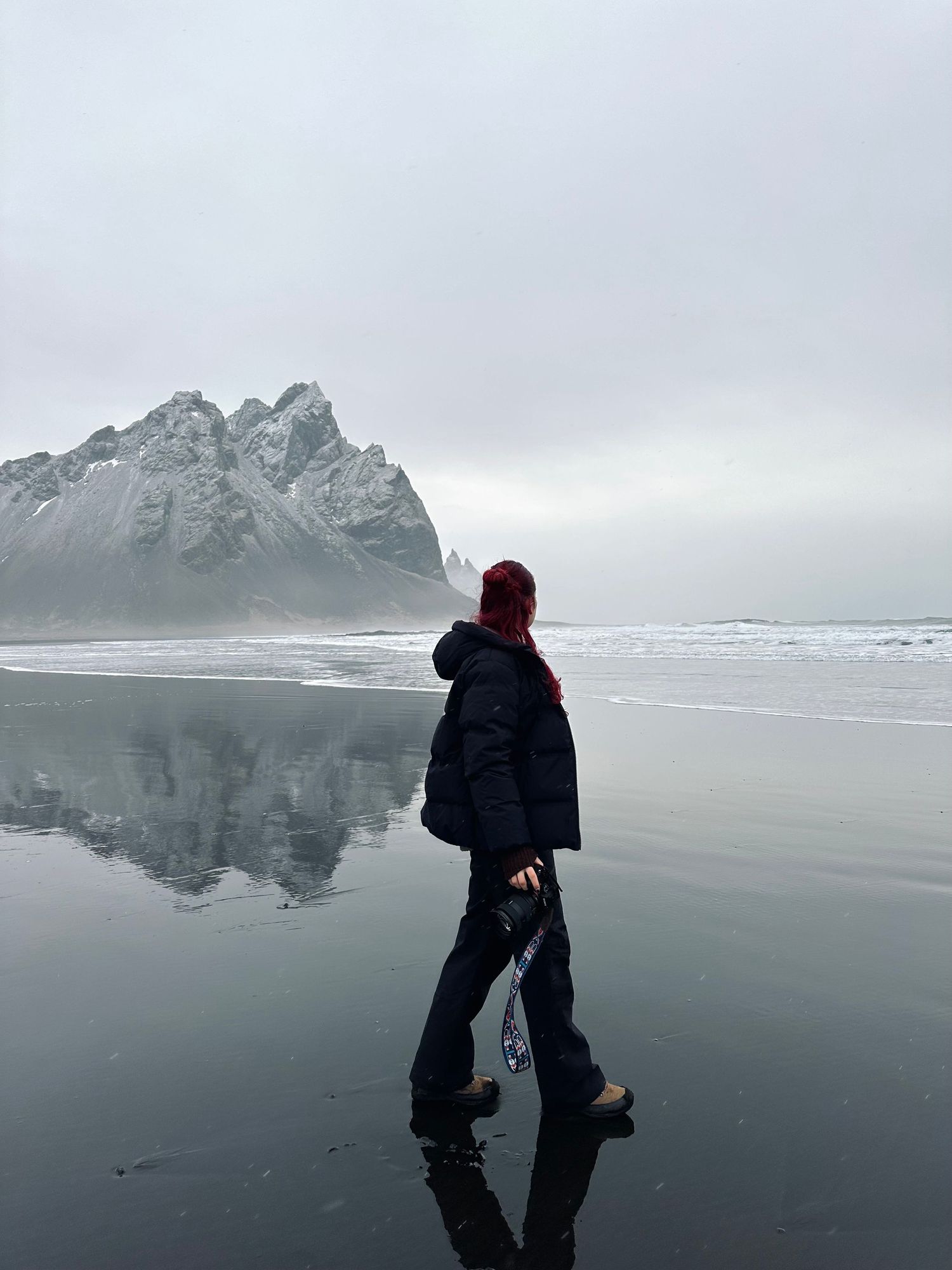 A solitary figure walks on a reflective black sand beach with dramatic mountain peaks in the misty background. Iceland.