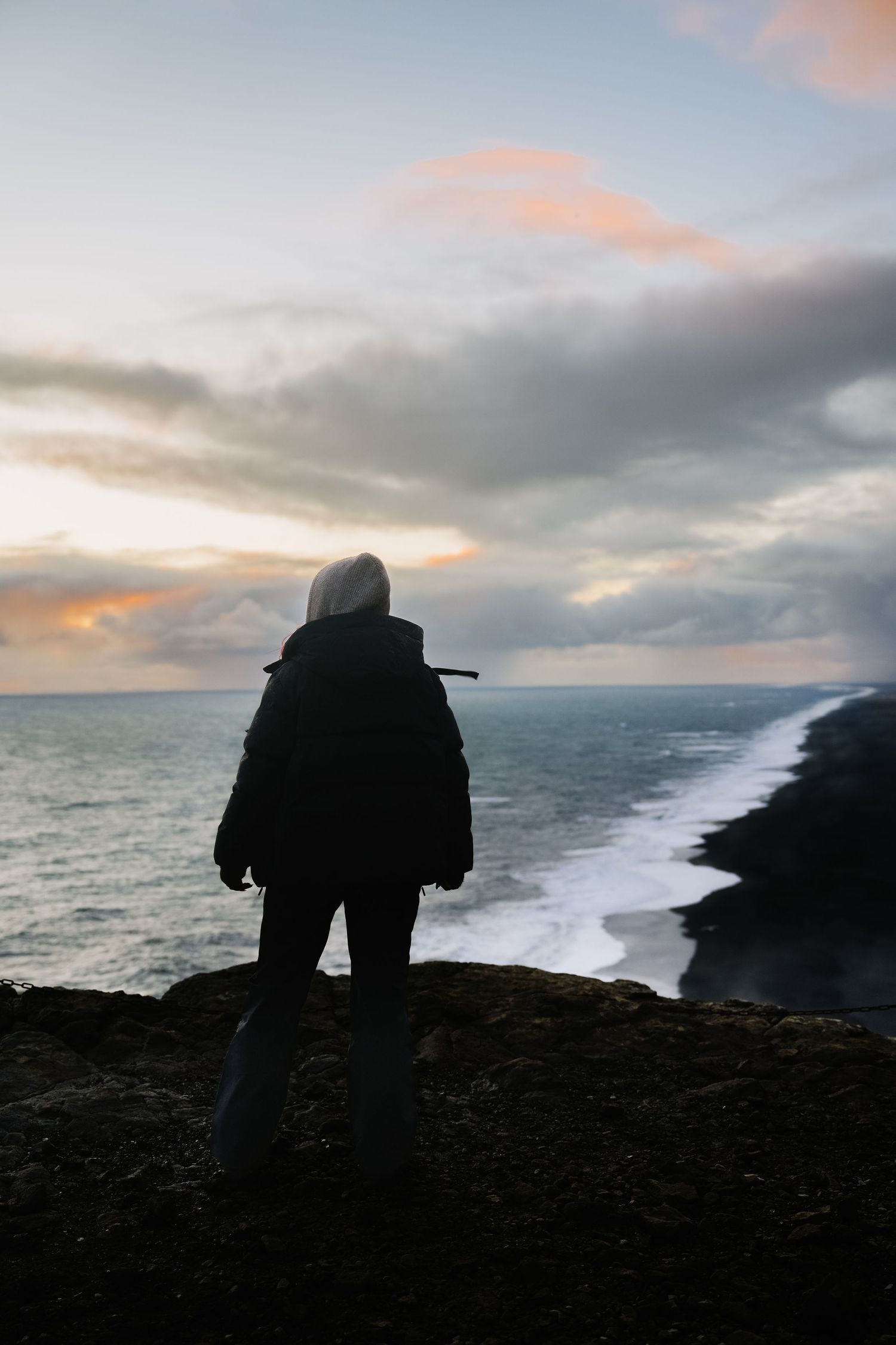 Silhouetted figures stand on rocky cliffs overlooking a dramatic ocean coastline at dusk. Iceland.