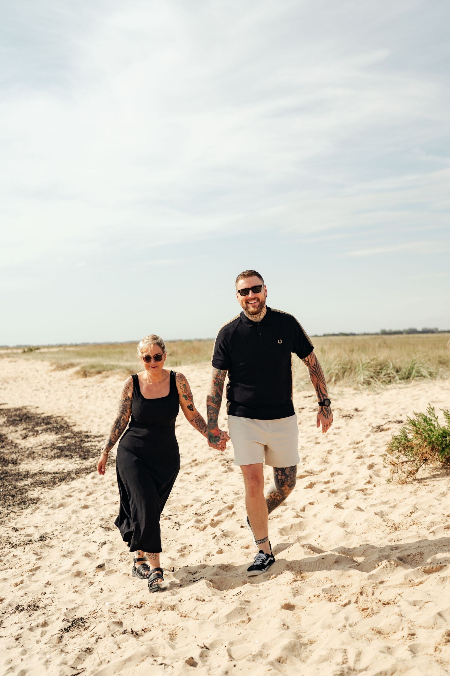 Couple walking hand in hand along sandy beach path in black and white outfits. Earlhams Beach, Harwich, Essex.