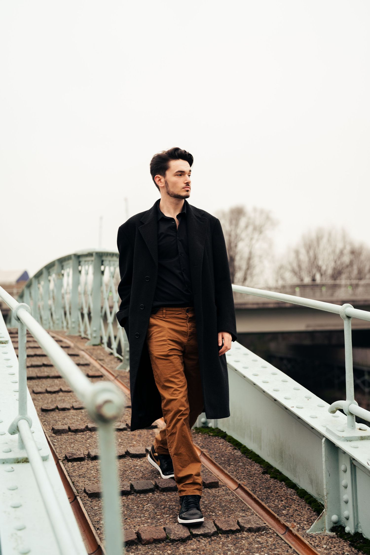 Figure in black coat and brown pants poses on white metal bleachers or stairs. London, UK.