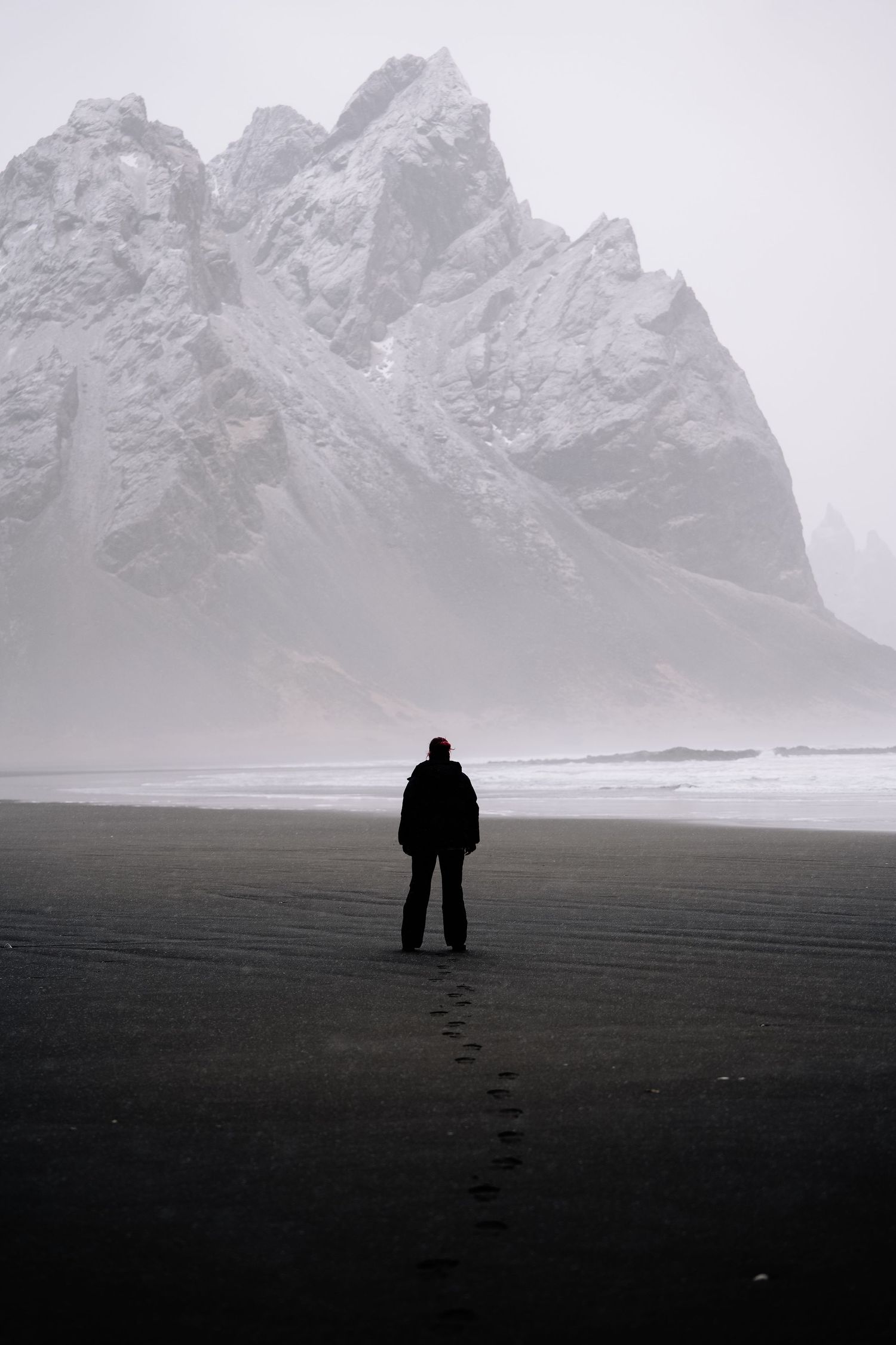 A solitary figure stands on a black sand beach with towering misty mountains in the background. Iceland.