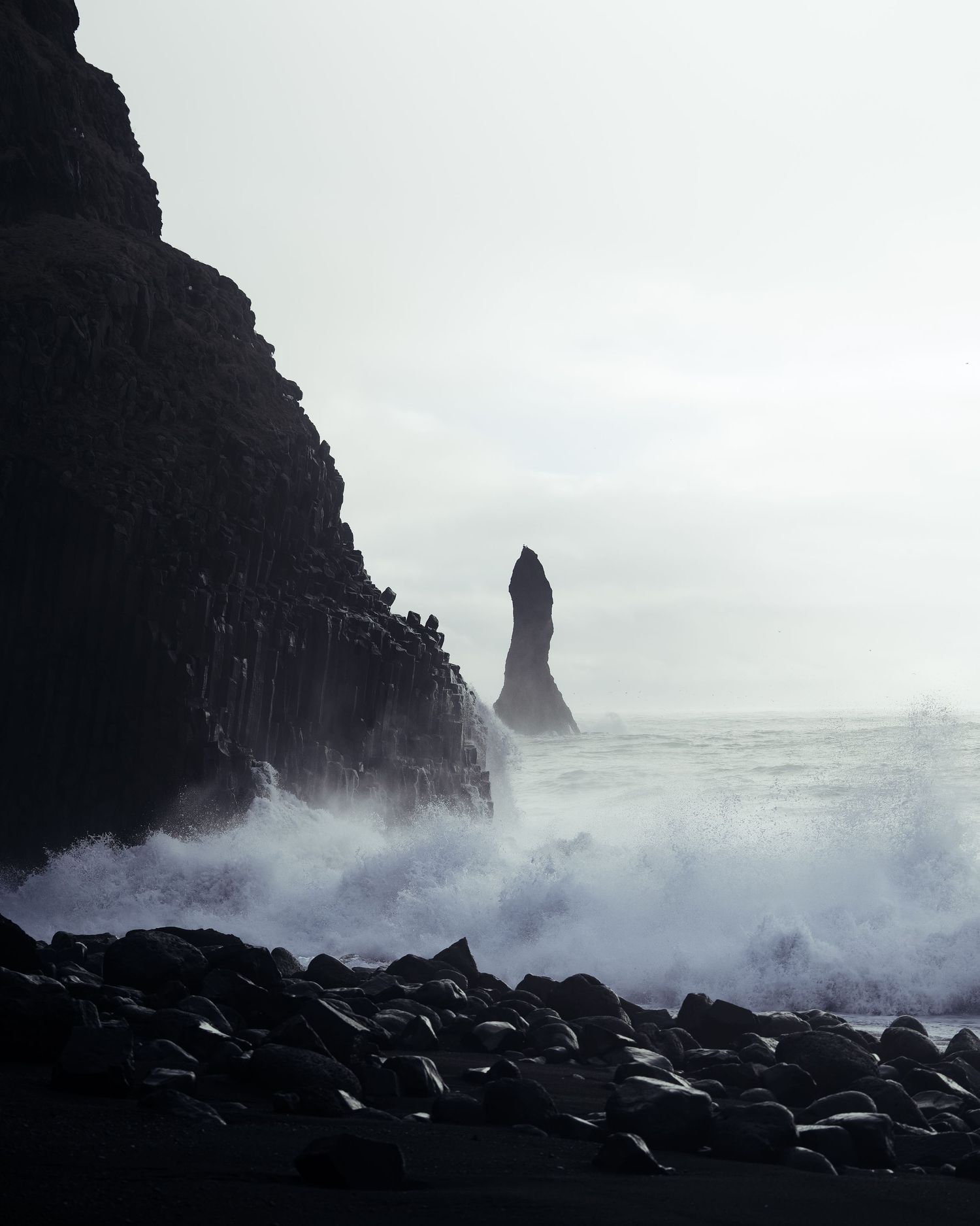 Dramatic black cliffs and sea stacks rise from churning waves in moody coastal scenery. Iceland.