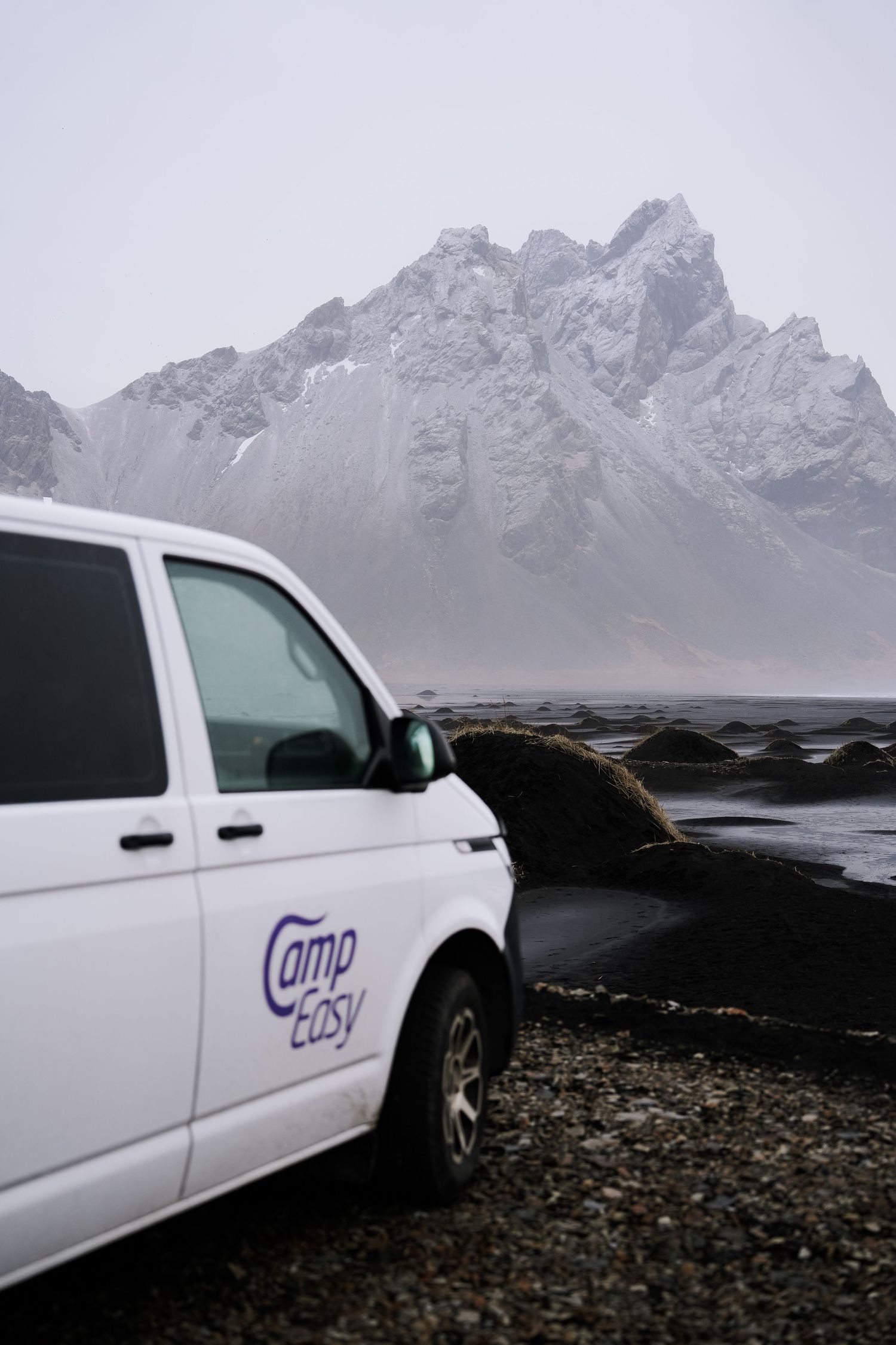 White company vans parked on a black sand beach with snow-covered mountains in the background. Iceland.