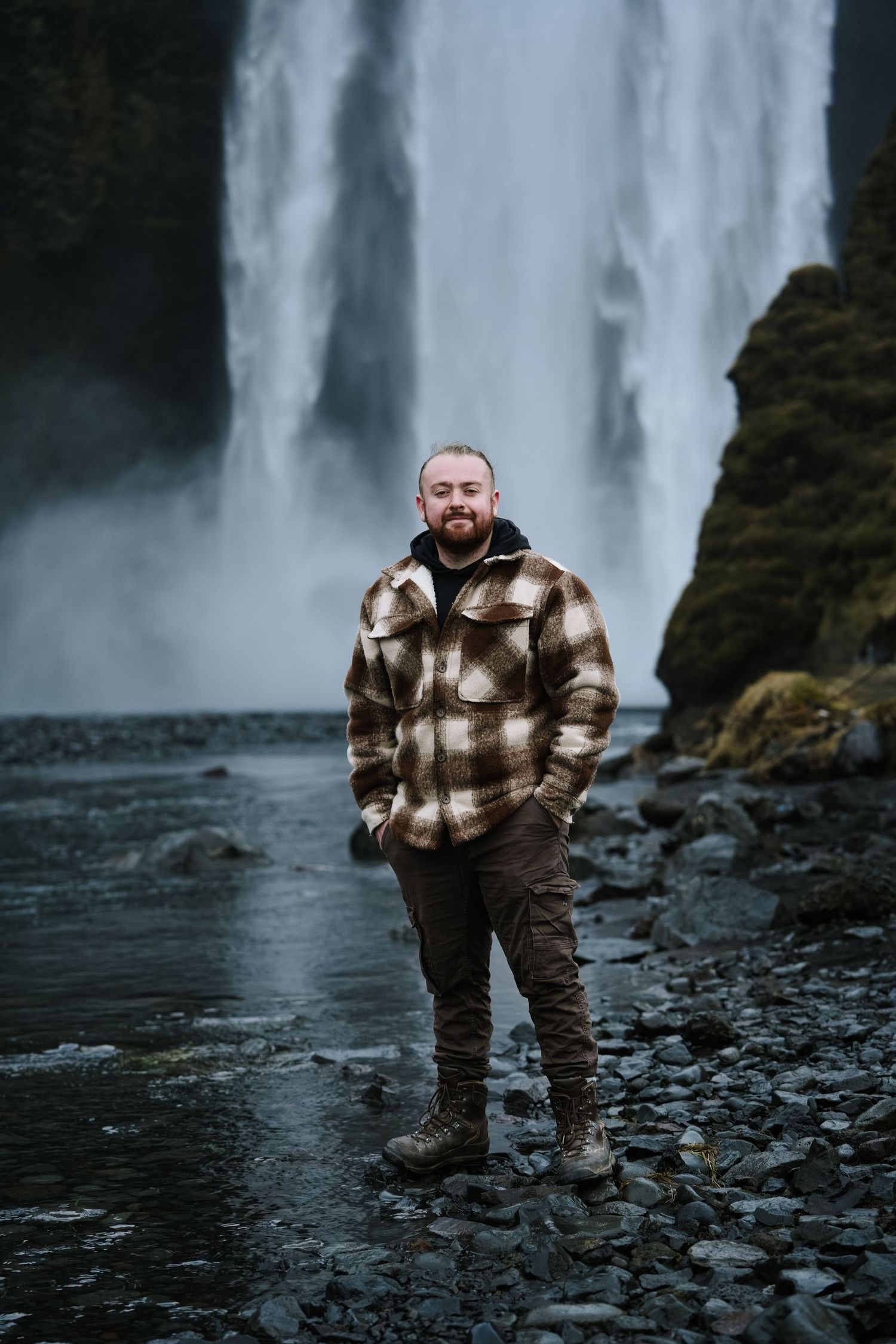 A person in a plaid jacket poses in front of a towering waterfall in Iceland.