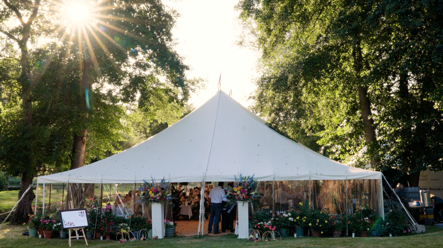 A panoramic view of an outdoor wedding venue with white tents and trees at sunset. Suffolk.