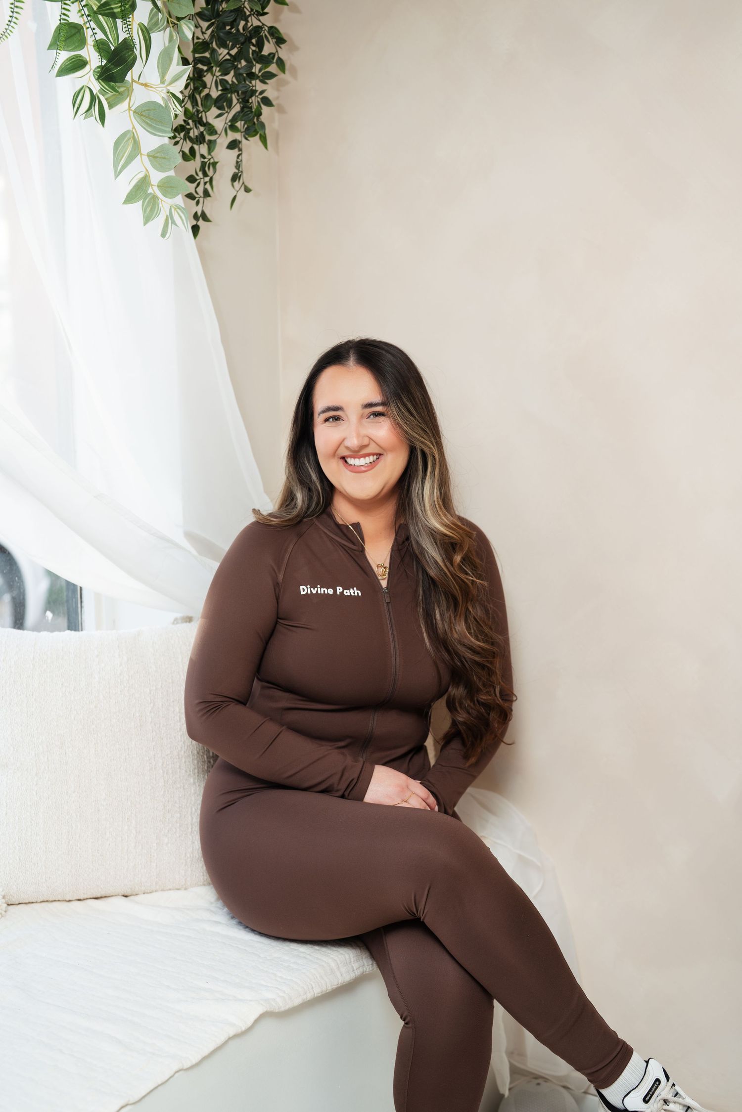 Person in brown loungewear poses comfortably on chair against white wall with hanging plants. Colchester, Essex.