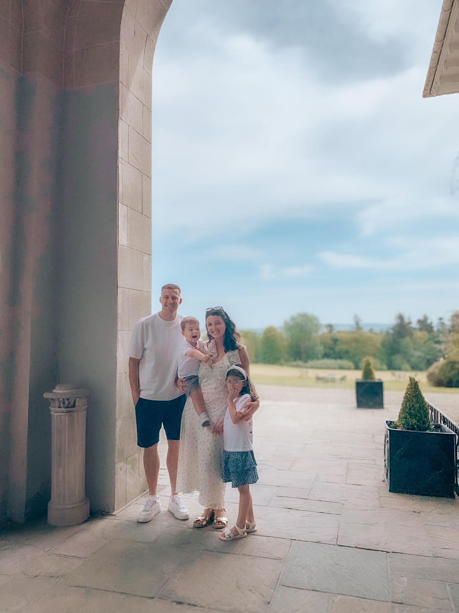 A family stands together under a stone archway on a sunny day at an outdoor location.