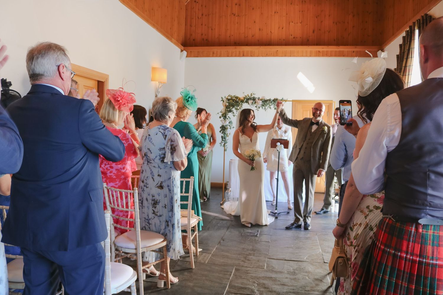 Wedding guests in colorful attire and Scottish kilts gather inside a rustic venue during a celebration ceremony.