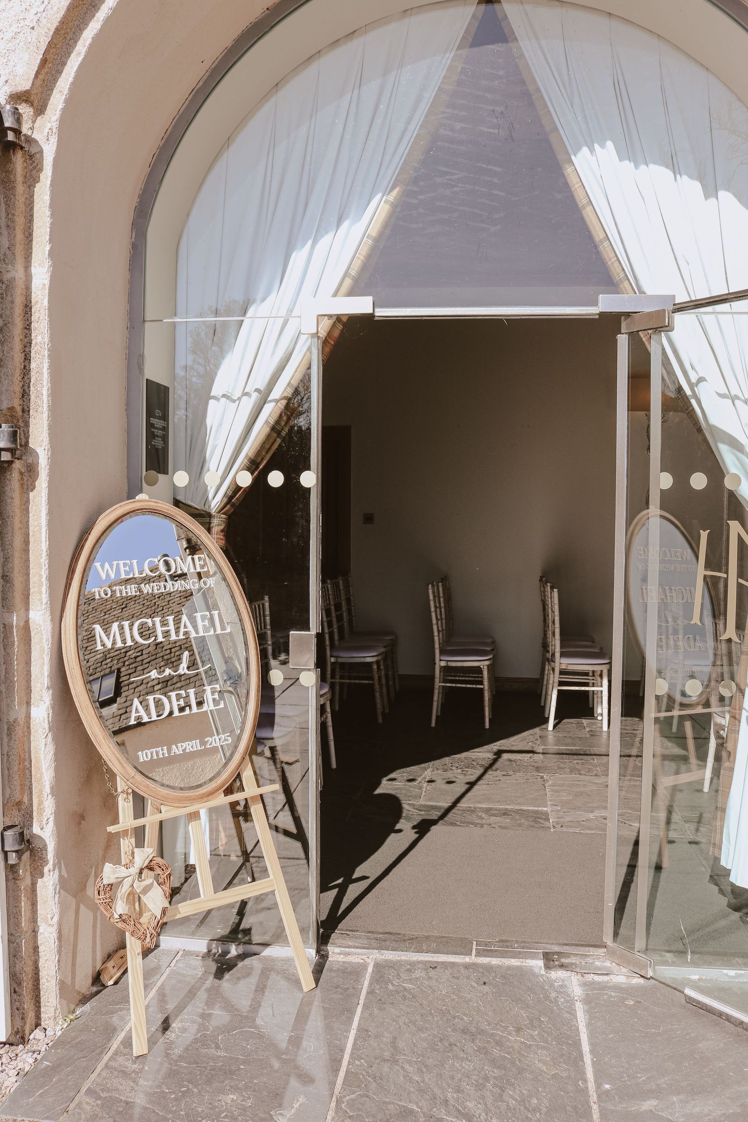 Rustic wooden welcome sign and white curtains frame an entrance to an indoor venue with chairs visible inside.