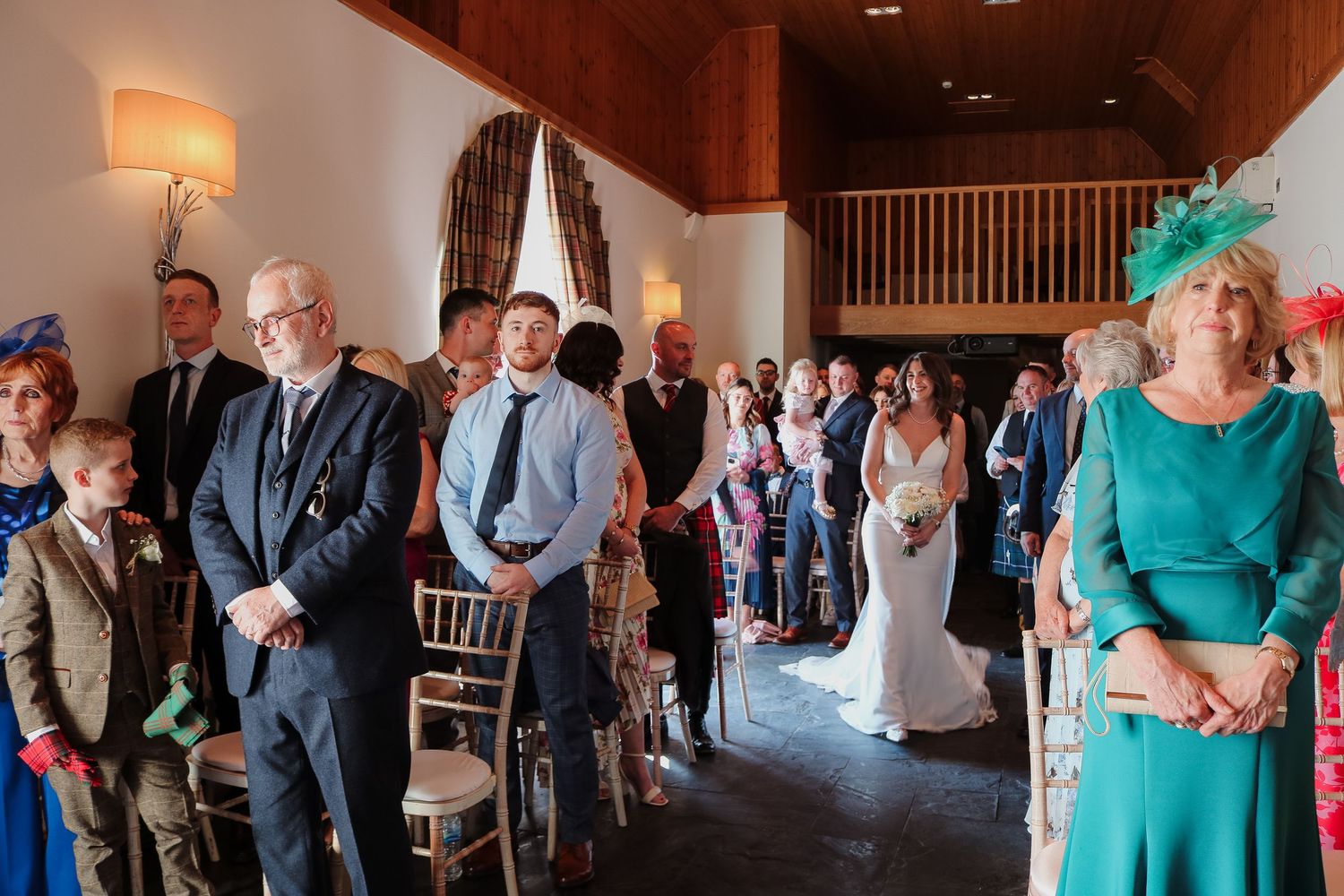 Wedding guests stand in a rustic indoor venue as they watch the ceremony proceedings from wooden chairs.