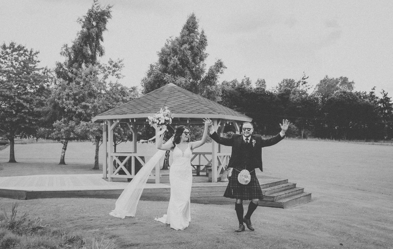 A vintage black and white photo shows a couple celebrating under a wooden gazebo in an outdoor garden setting.