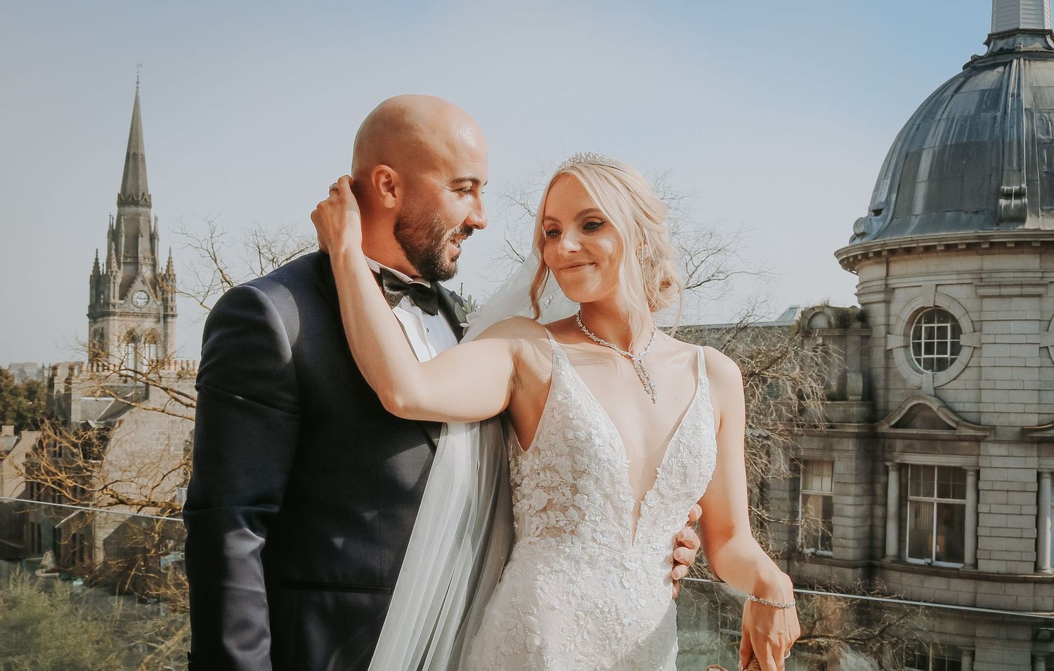 A couple shares a romantic moment in wedding attire with a beautiful church spire and domed building in the background.