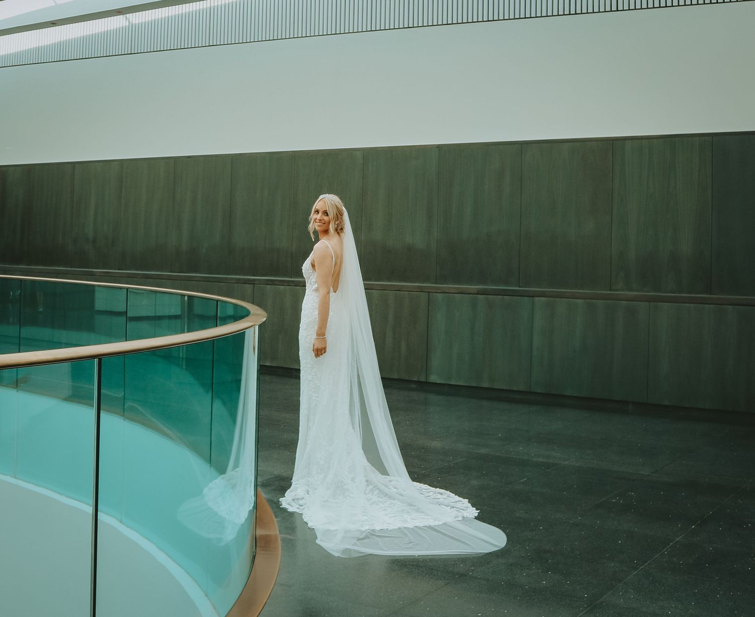 A bride in a flowing white gown and veil poses along a curved modern architectural walkway.