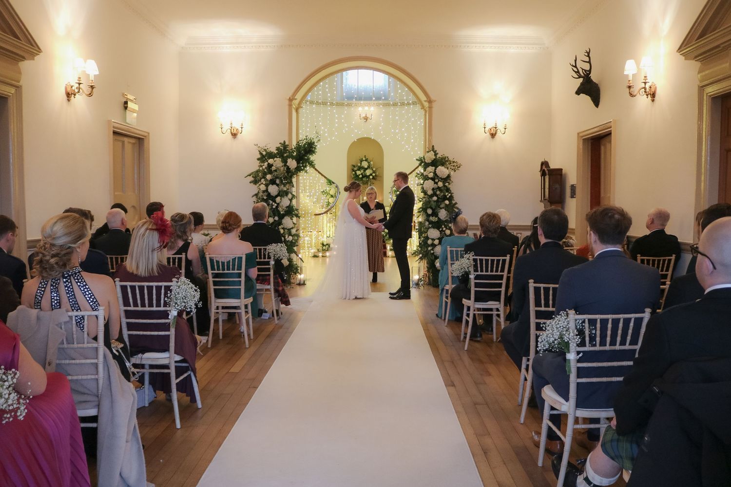 Indoor wedding ceremony with white runner and rows of silver chairs in an elegant venue lit by wall sconces.