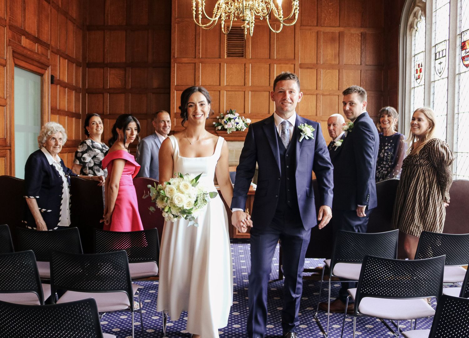 Wedding guests mingle and socialize in an elegant ballroom with a chandelier and large windows.