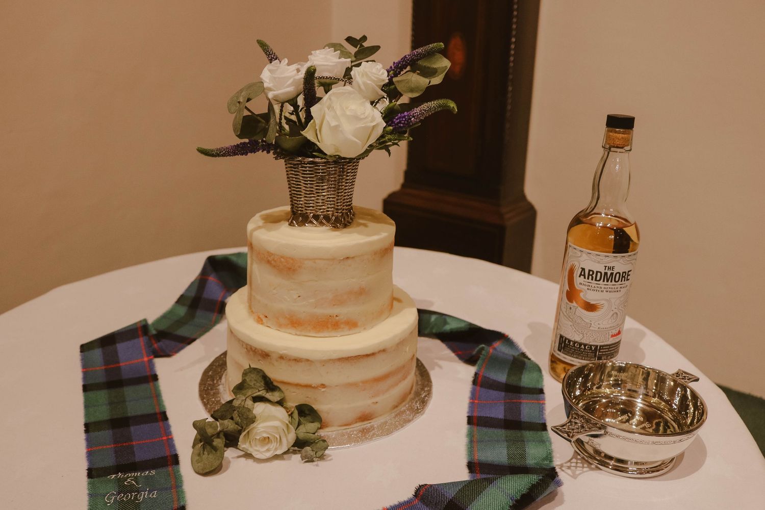 Semi-naked wedding cake displayed with white roses and greenery on a plaid cloth next to a whiskey bottle.