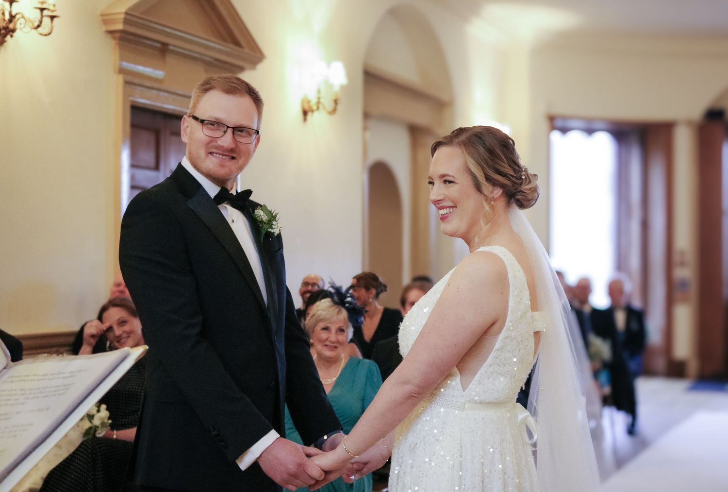A couple exchanges vows during their indoor wedding ceremony as guests look on in an elegant venue with warm lighting.