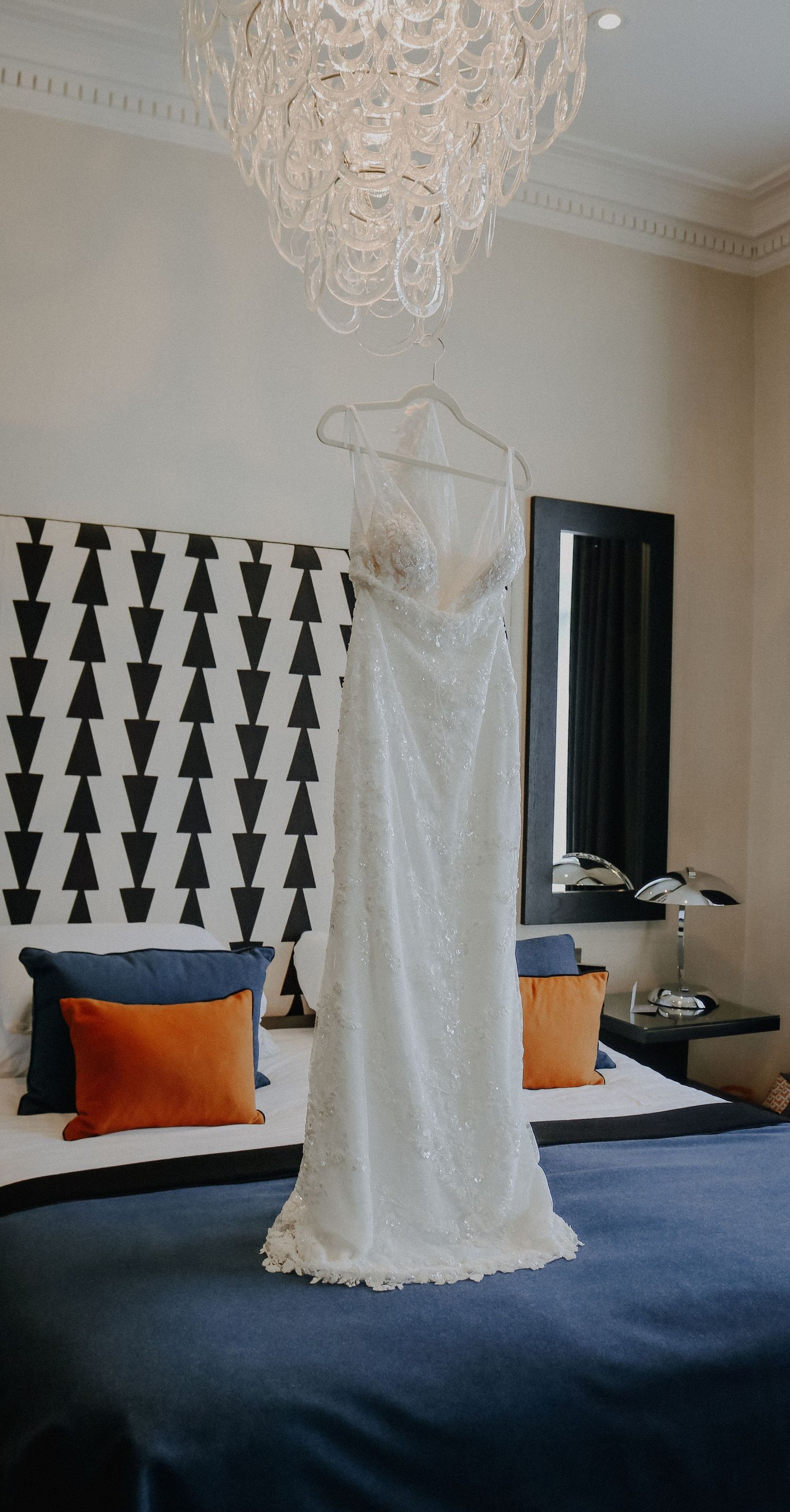 An elegant white lace wedding dress hangs in a modern bedroom with geometric black and white wall art and orange pillows.