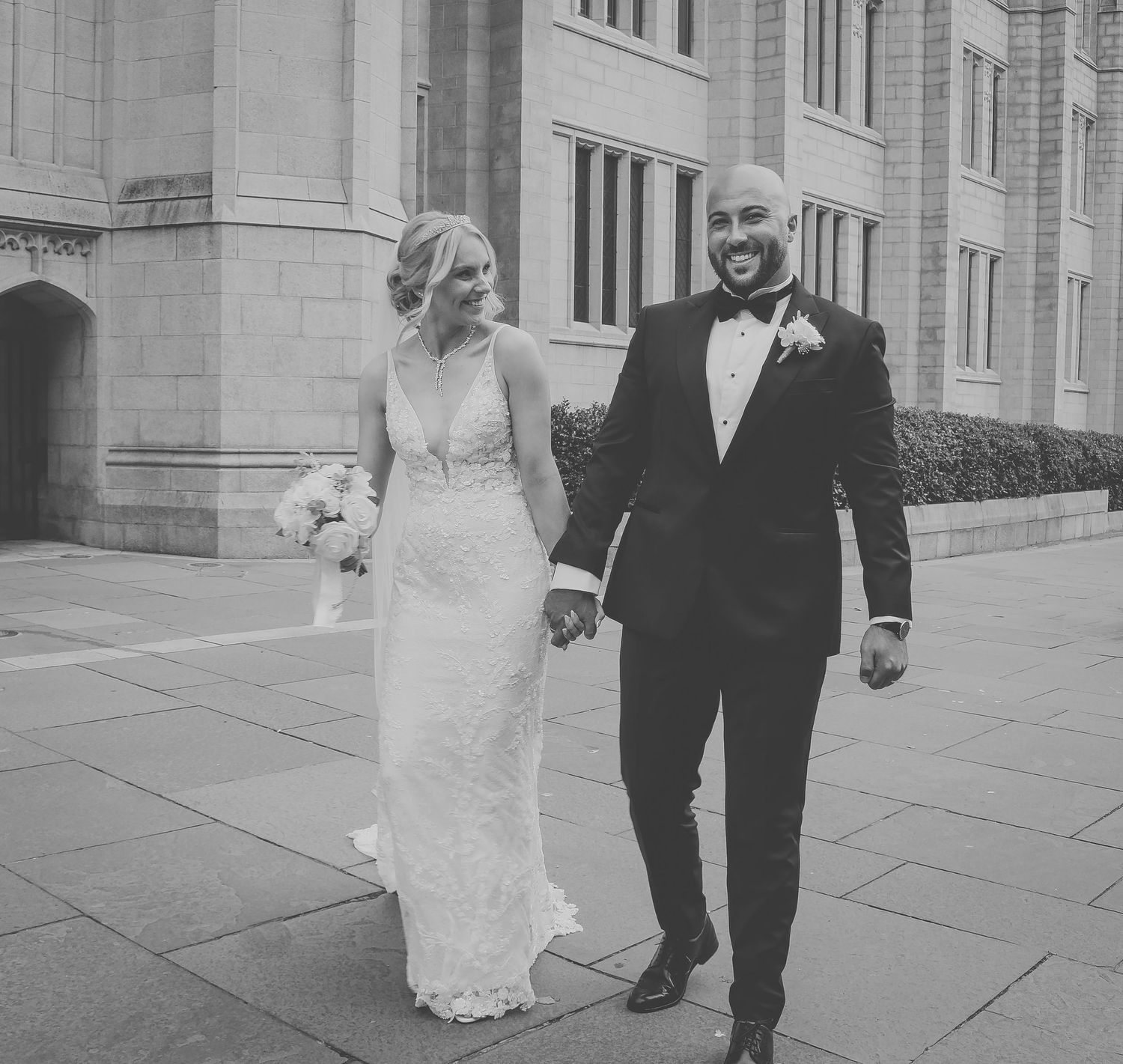 A couple walks hand in hand outside a grand stone building in black and white wedding photography.