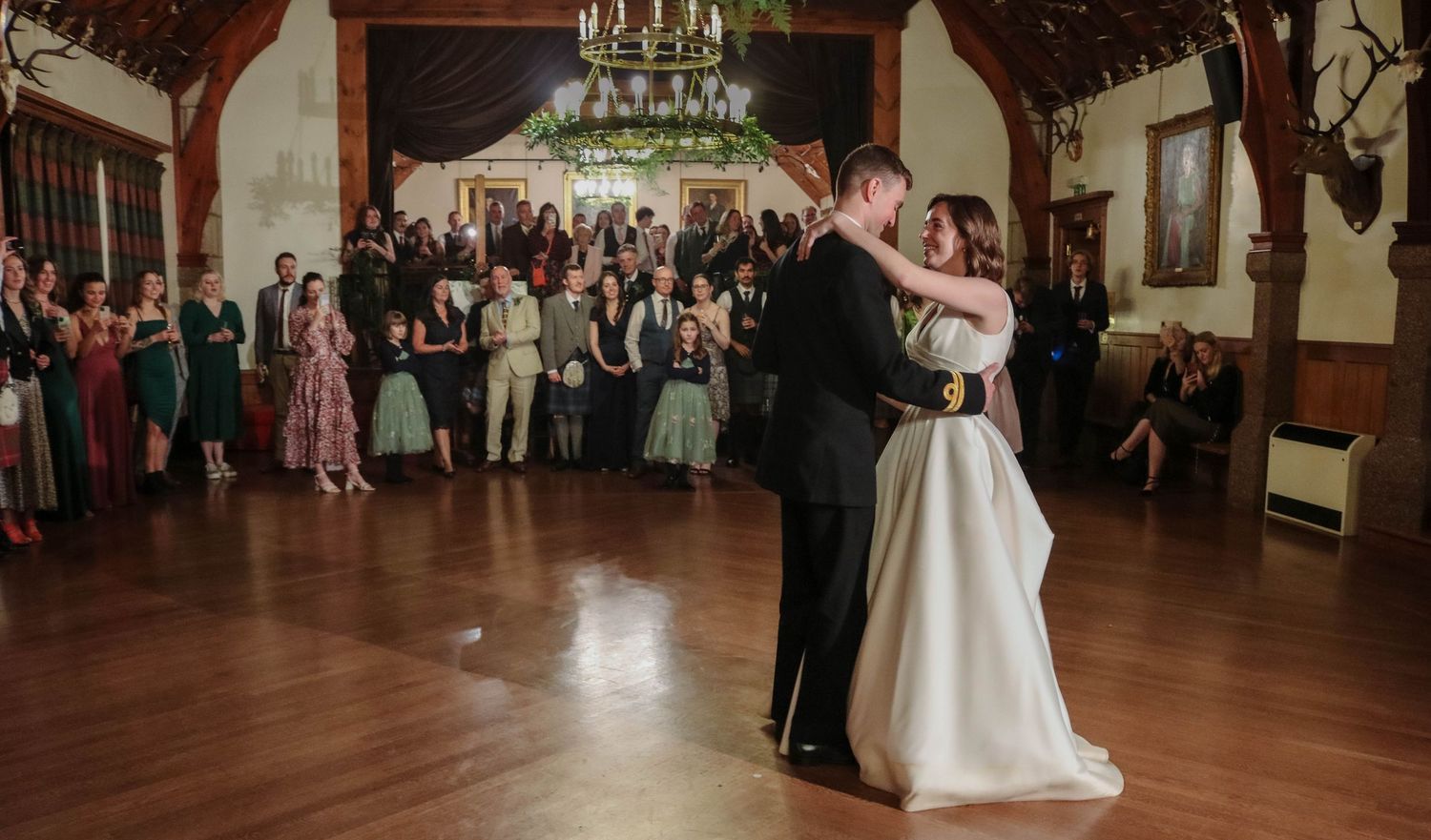 A couple shares their first dance in an elegant ballroom with wood paneling and chandeliers while guests watch from the sides.