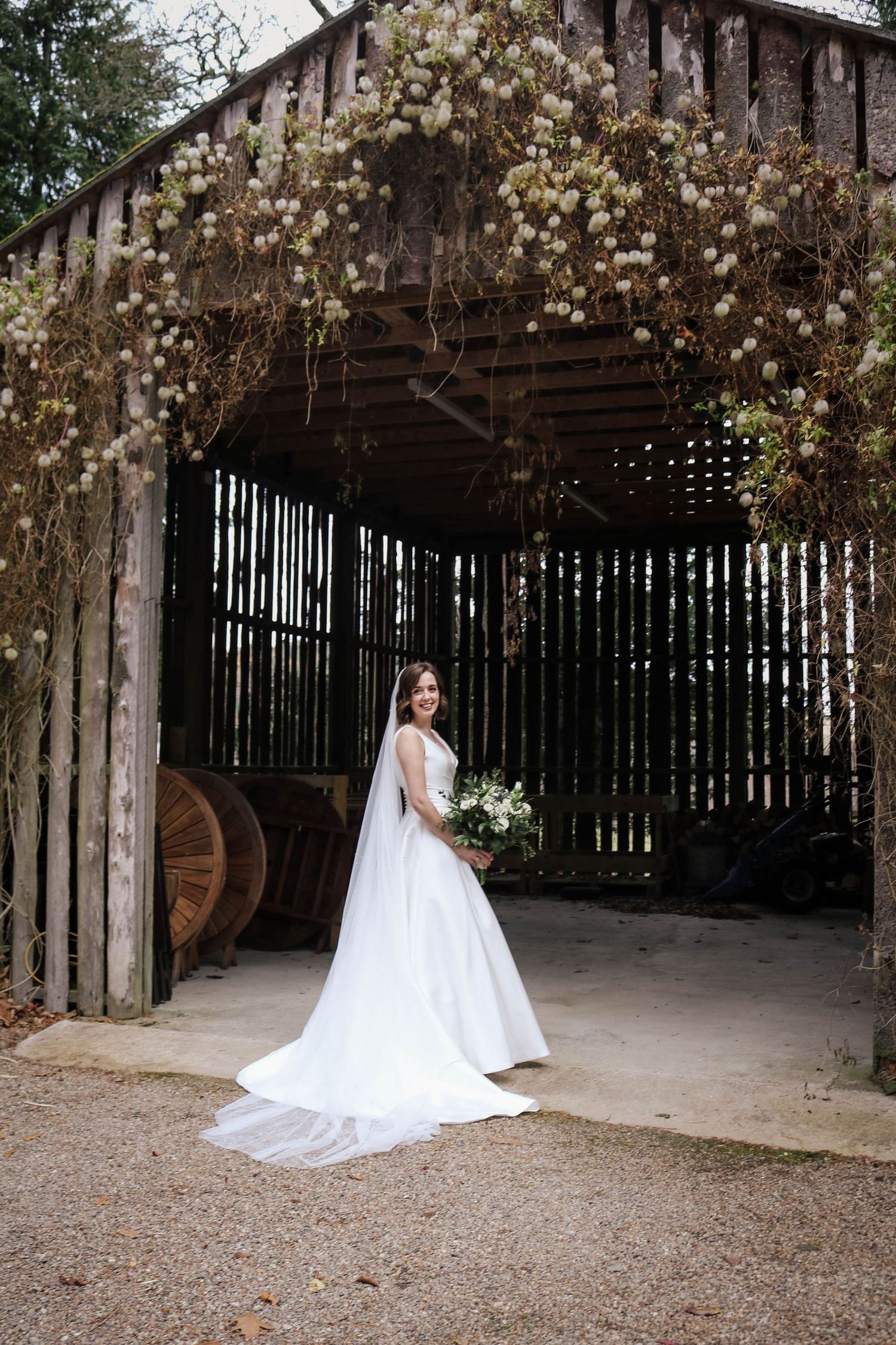 Rustic barn venue decorated with cascading white flowers creates romantic backdrop for elegant bridal portrait.