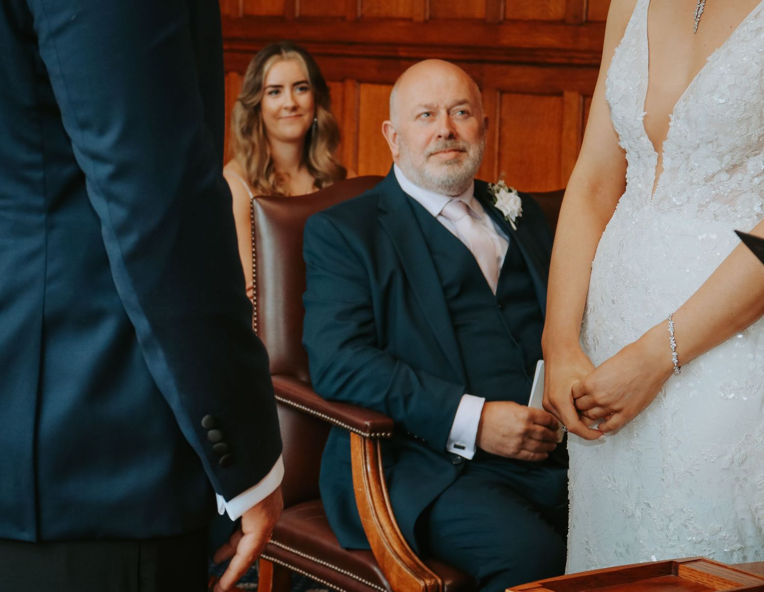 Wedding ceremony participants seated in elegant wooden chairs with rich wood paneling in background.