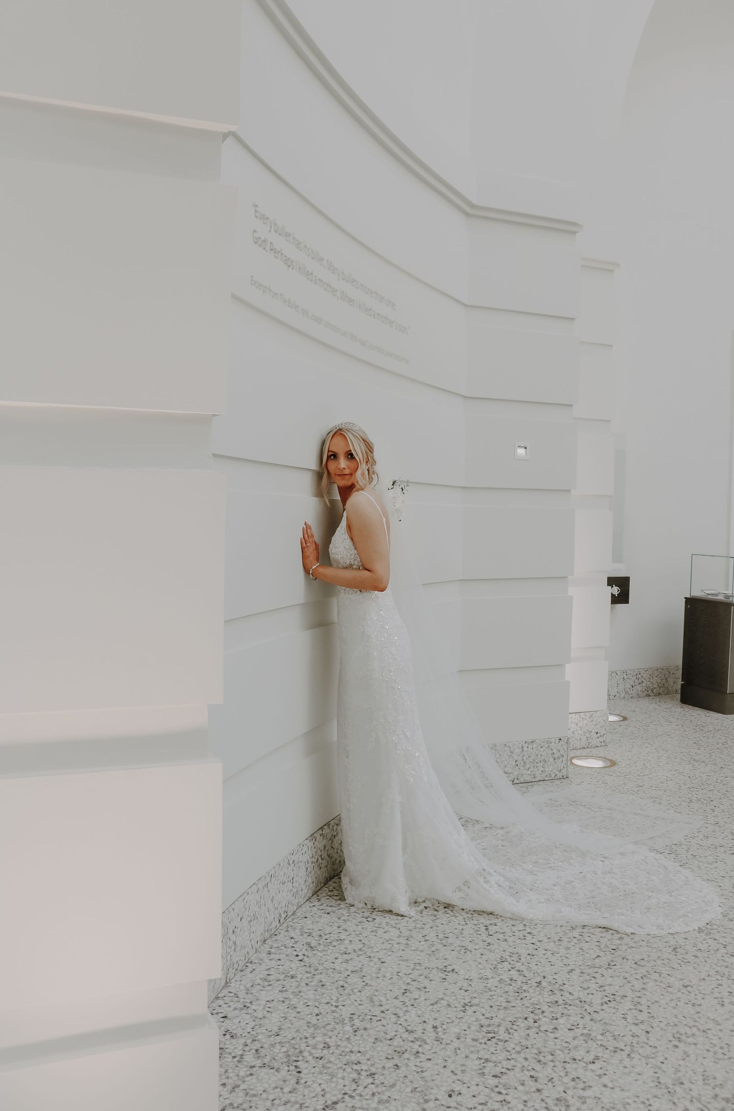 A bride in a long white gown poses against a crisp white wall.