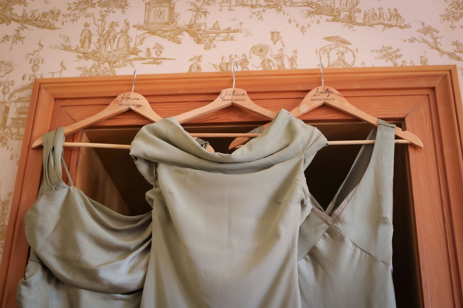 Three sage green bridesmaid dresses hanging on wooden hangers against a blush pink wall with toile pattern wallpaper.