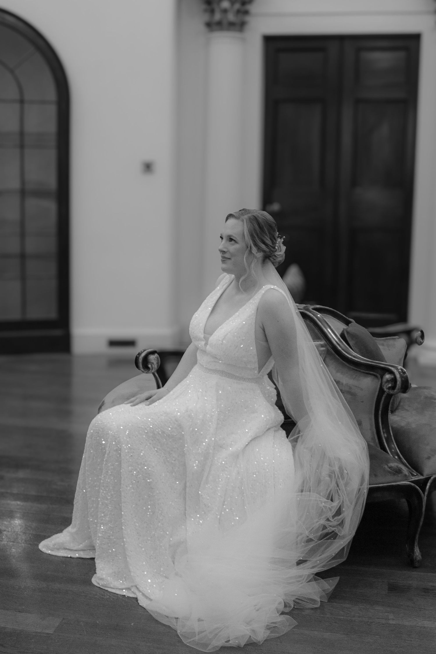 Black and white portrait of a bride in a sparkling V-neck wedding gown sitting elegantly on an antique bench.